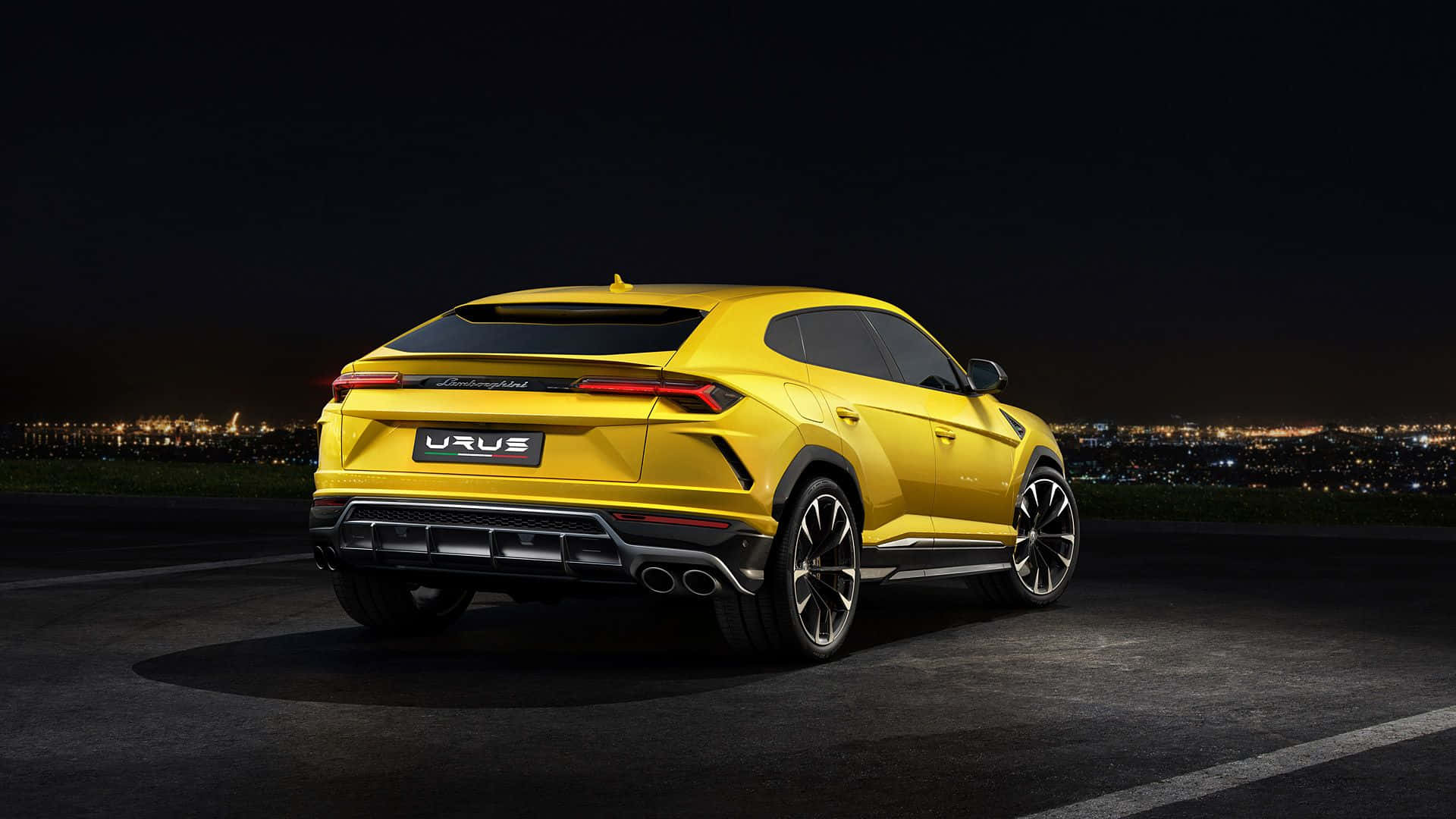 Lamborghini Urus 桌布 壁紙 — 一輛鮮黃色的藍寶堅尼 Urus SUV 在夜間停泊，遠處城市燈光閃爍。