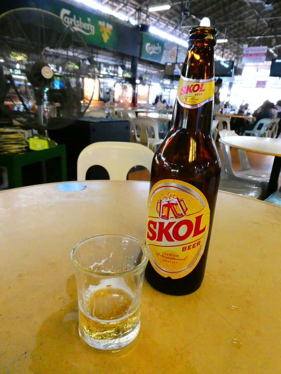  壁紙 — 一個棕色Skol啤酒瓶和半滿的玻璃杯放在黃色桌子上，室內餐廳內有風扇和顧客，氣氛熱鬧。
