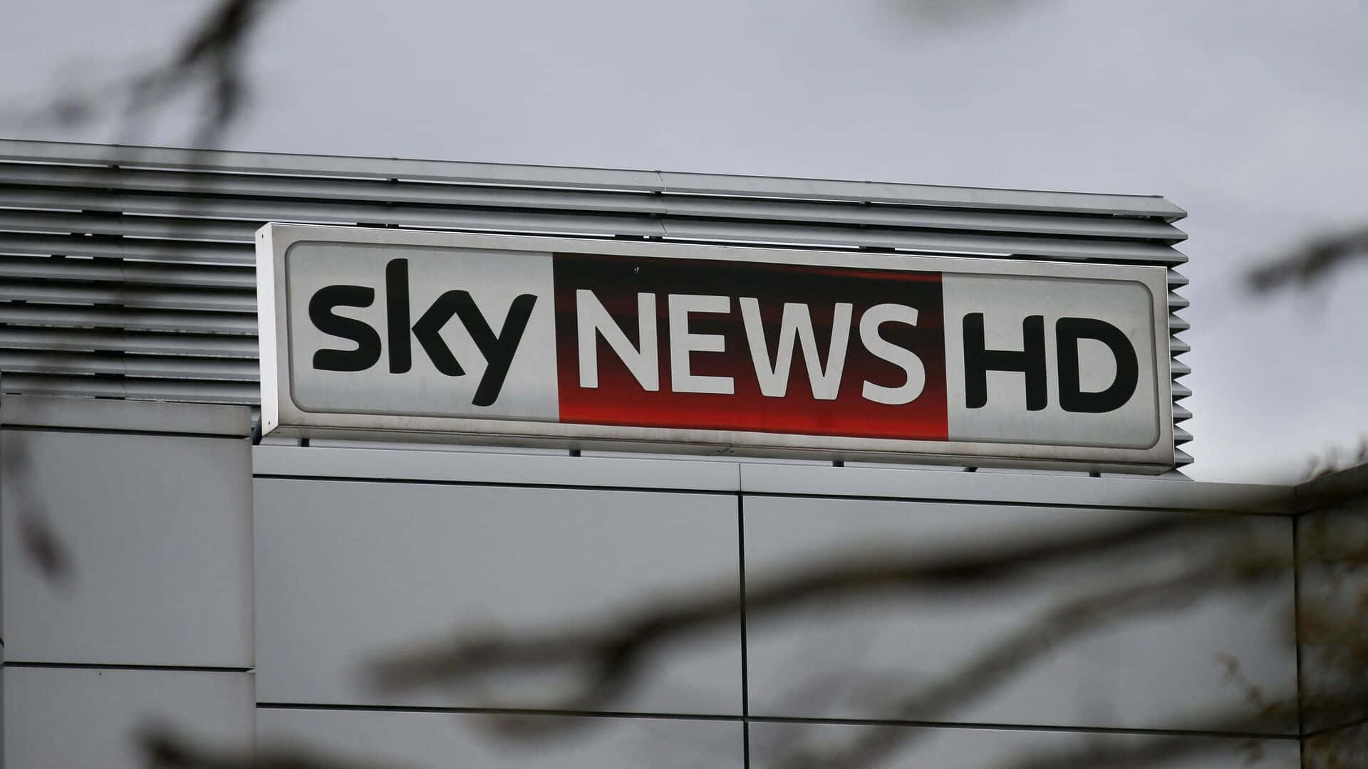 高清 桌布 壁紙 — Sky News HD 標誌的特寫，安裝在現代建築物上，前景有模糊的樹枝。