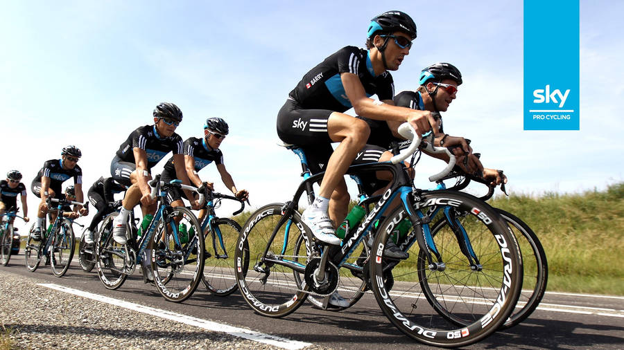 桌布 壁紙 — Sky Pro Cycling 車隊的專業自行車手在陽光明媚的道路上以緊密隊形比賽，背景是綠色山丘。