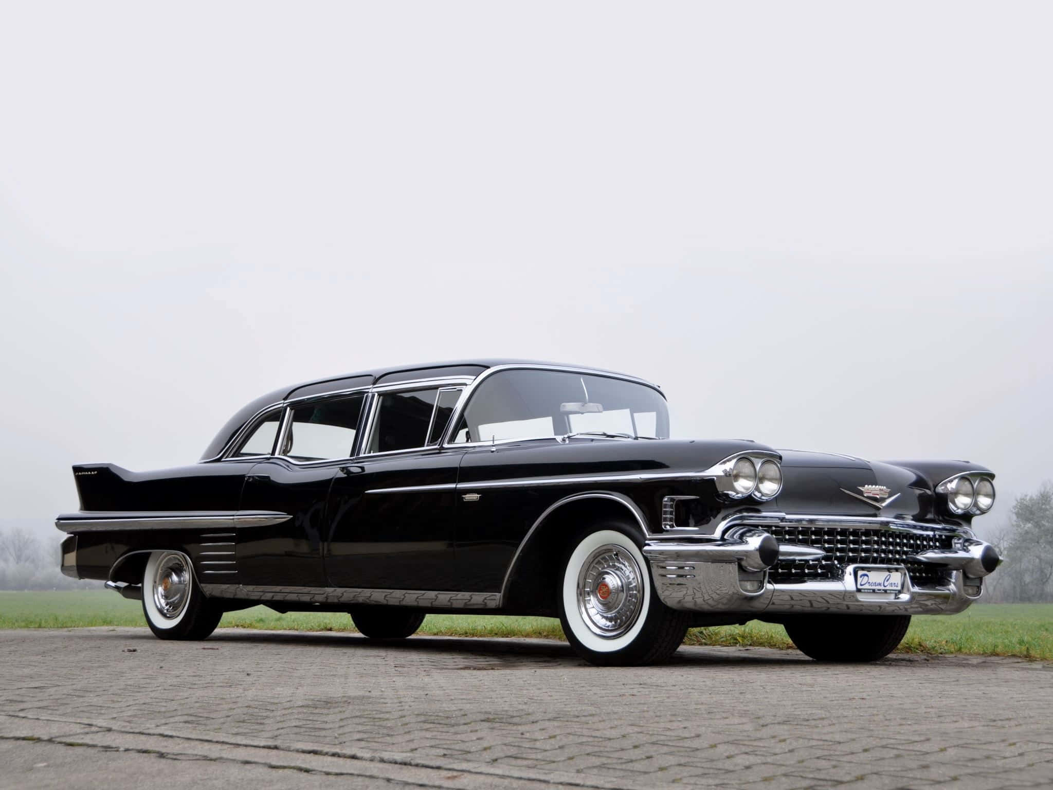Cadillac Fleetwood 桌布 壁紙 — 一輛時尚的黑色1959年凱迪拉克，帶有鍍鉻裝飾和白色輪胎，停在鵝卵石路面上，天空多雲。