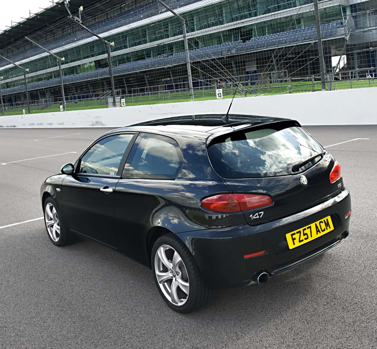 Alfa Romeo 147 桌布 壁紙 — 一輛時尚的黑色愛快羅密歐147掀背車停在瀝青賽車場上，背景有看台。