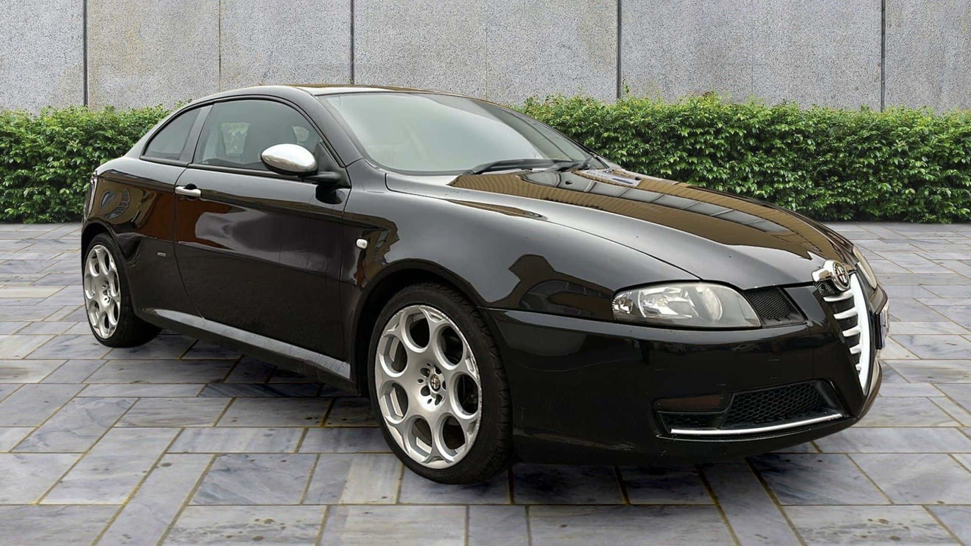 Alfa Romeo Gt 桌布 壁紙 — 一輛時尚的黑色愛快羅密歐雙門跑車在日光下閃耀，停放在由綠色樹籬環繞的石頭庭院上。