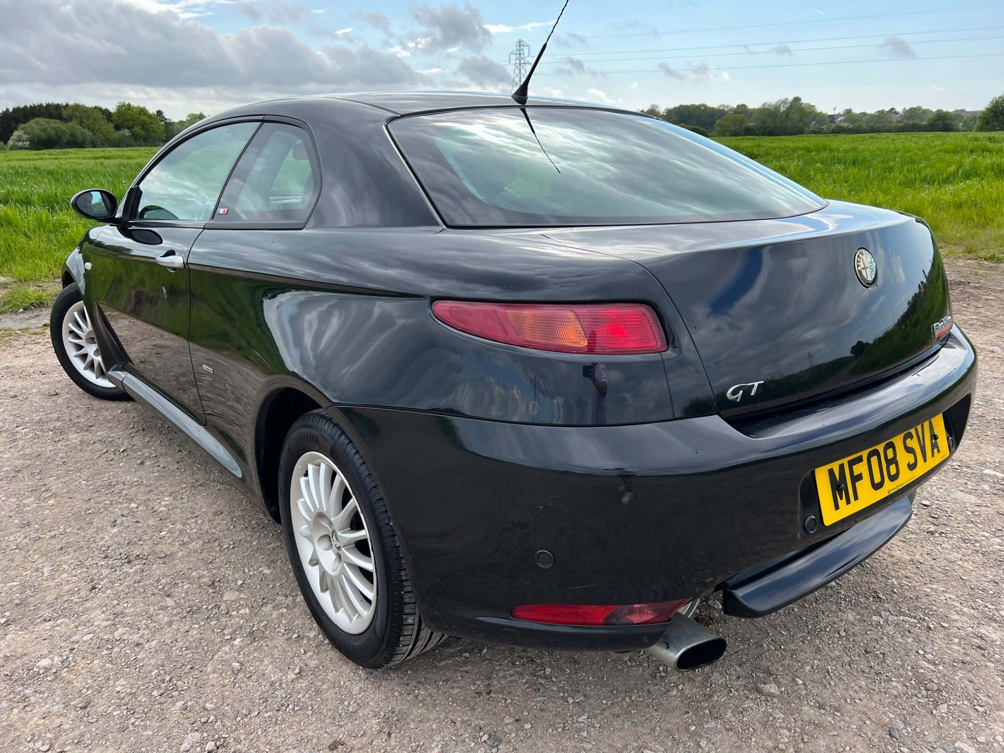 Alfa Romeo Gt 桌布 壁紙 — 一輛時尚的黑色愛快羅密歐GT雙門轎跑車停在碎石路上，背景是綠色的田野和多雲的天空。
