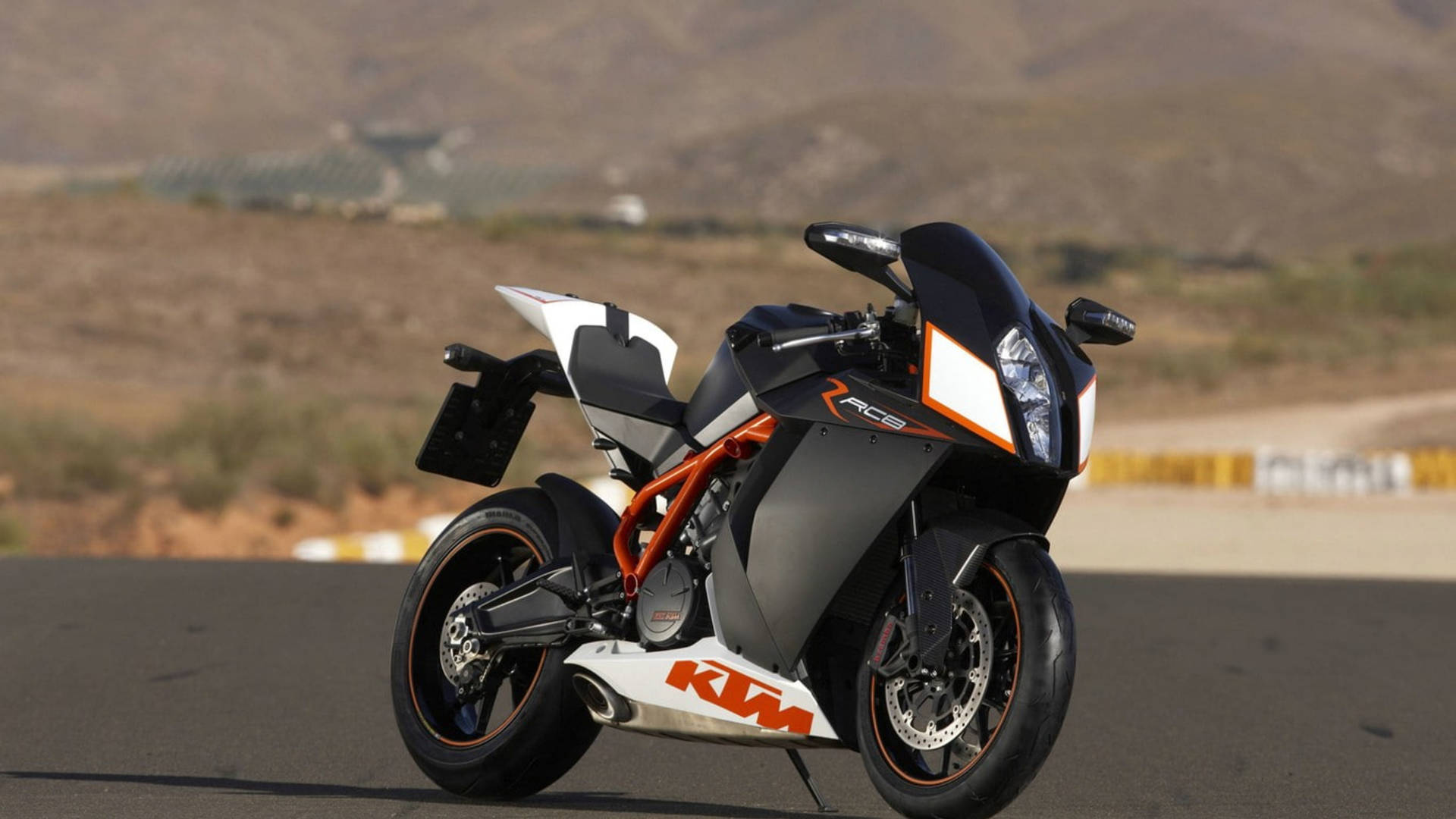 黑色和橙色KTM RC8摩托車在賽車場上，後面有沙漠山丘。 — 4K 壁紙