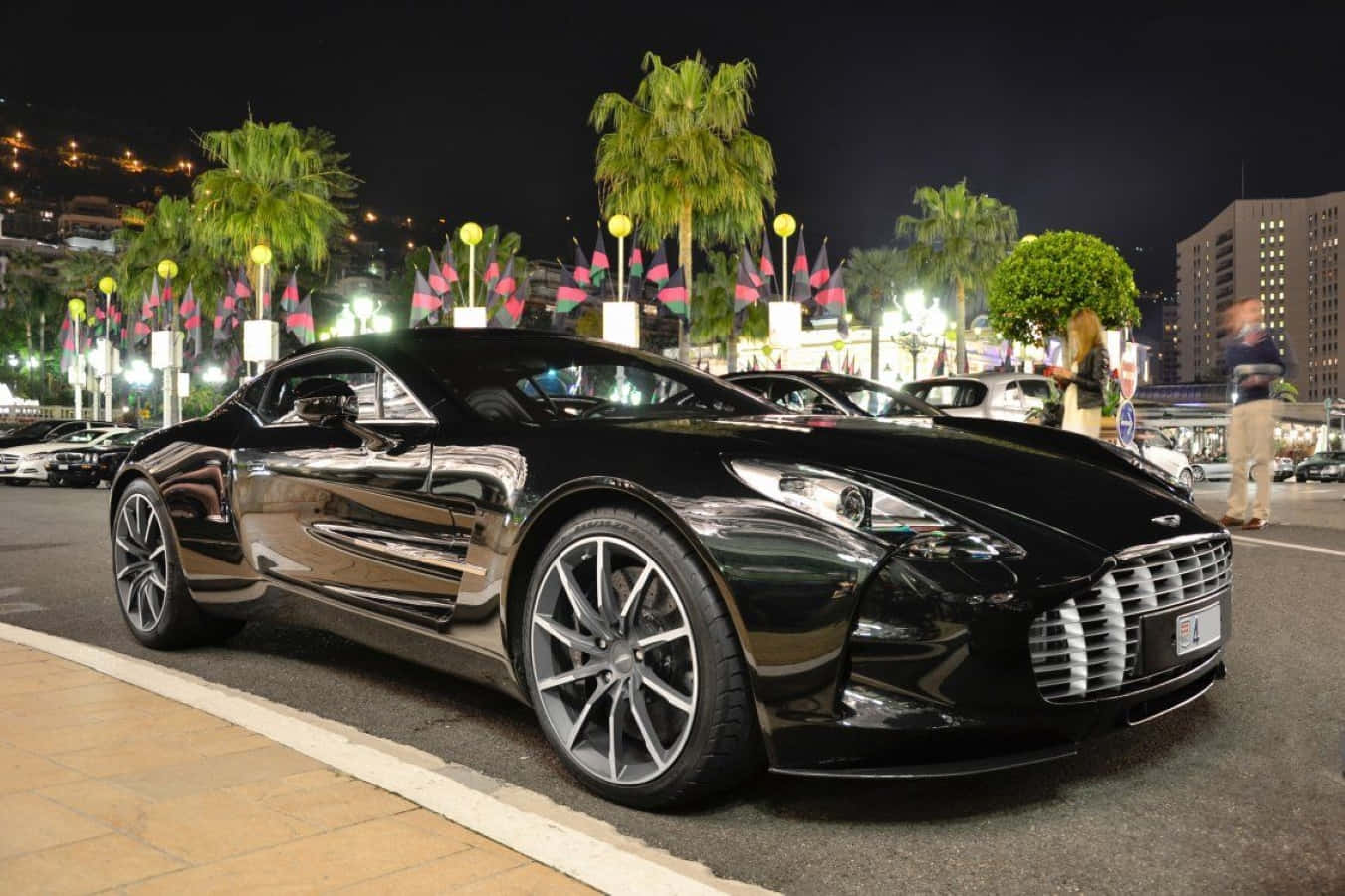 Aston Martin One-77 桌布 壁紙 — 一輛時尚的黑色Aston Martin跑車在夜燈下閃閃發光，位於棕櫚樹成蔭的街道上，行人模糊。