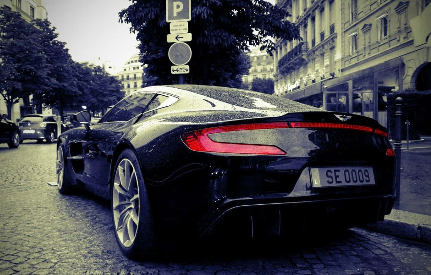 Aston Martin One-77 桌布 壁紙 — 一輛時尚的黑色Aston Martin跑車停在濕漉漉的鵝卵石街道上，背景是復古歐洲建築。