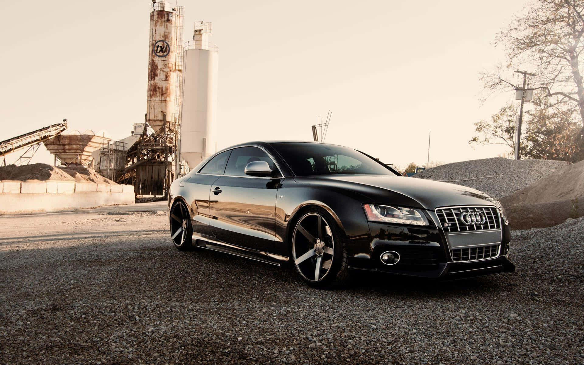 Audi S5 桌布 壁紙 — 一輛時尚的黑色奧迪轎跑車在溫暖的陽光下閃閃發光，停在工業筒倉和光禿樹木附近的礫石上。