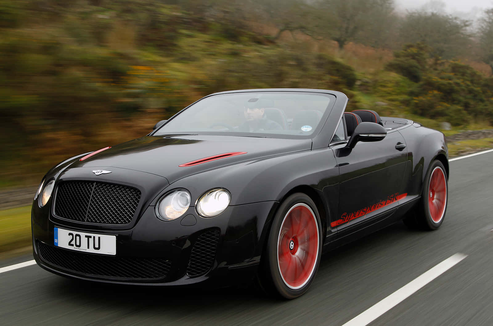 Bentley Continental Supersports 桌布 壁紙 — 一輛時尚的黑色賓利敞篷車帶有紅色點綴，在模糊的綠色風景中沿著蜿蜒的道路疾馳。
