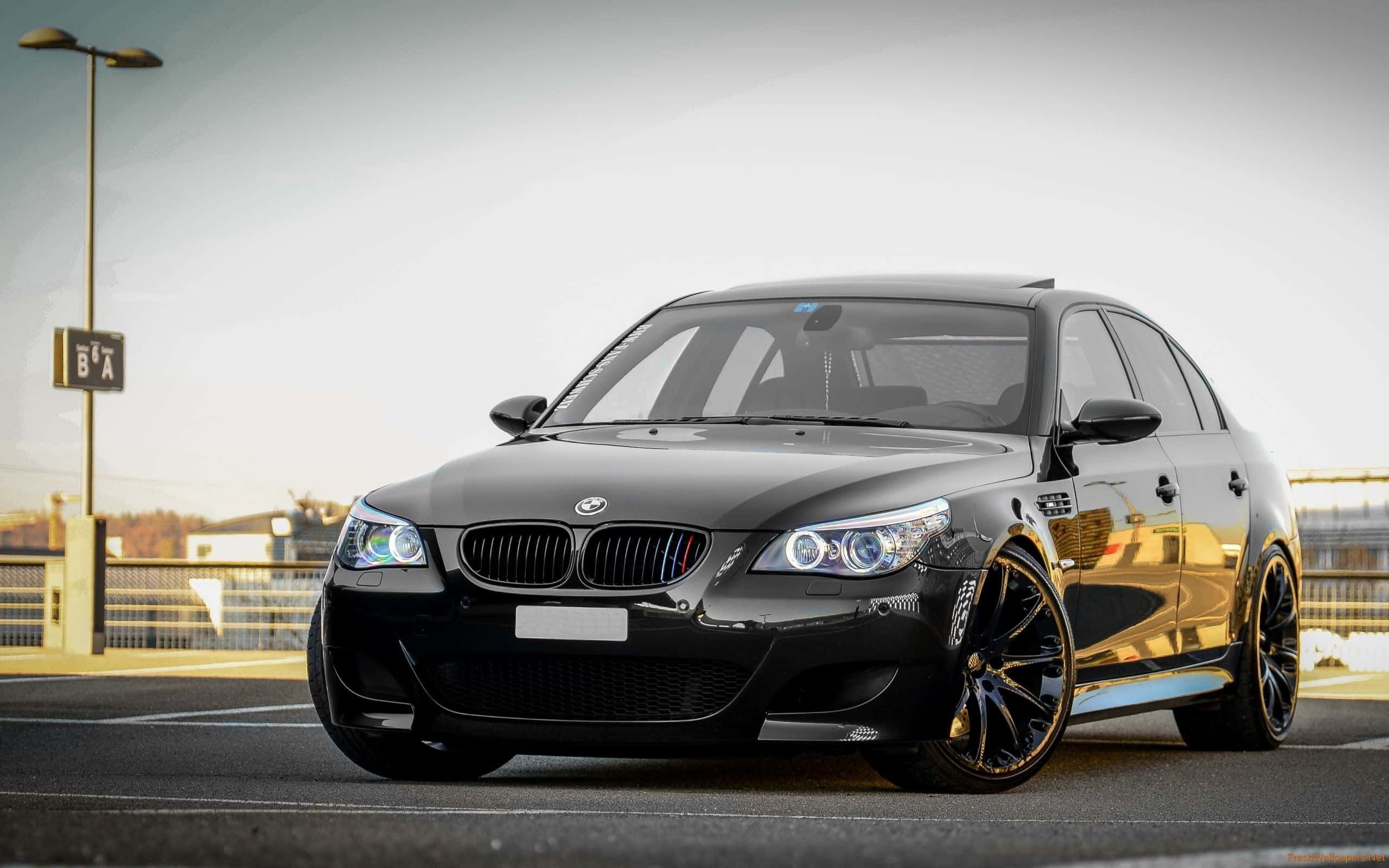 Bmw M5 桌布 壁紙 — 一輛時尚的黑色BMW M5運動轎車停在瀝青停車場上，車頭燈閃爍，配備定制輪圈，在柔和日光下。