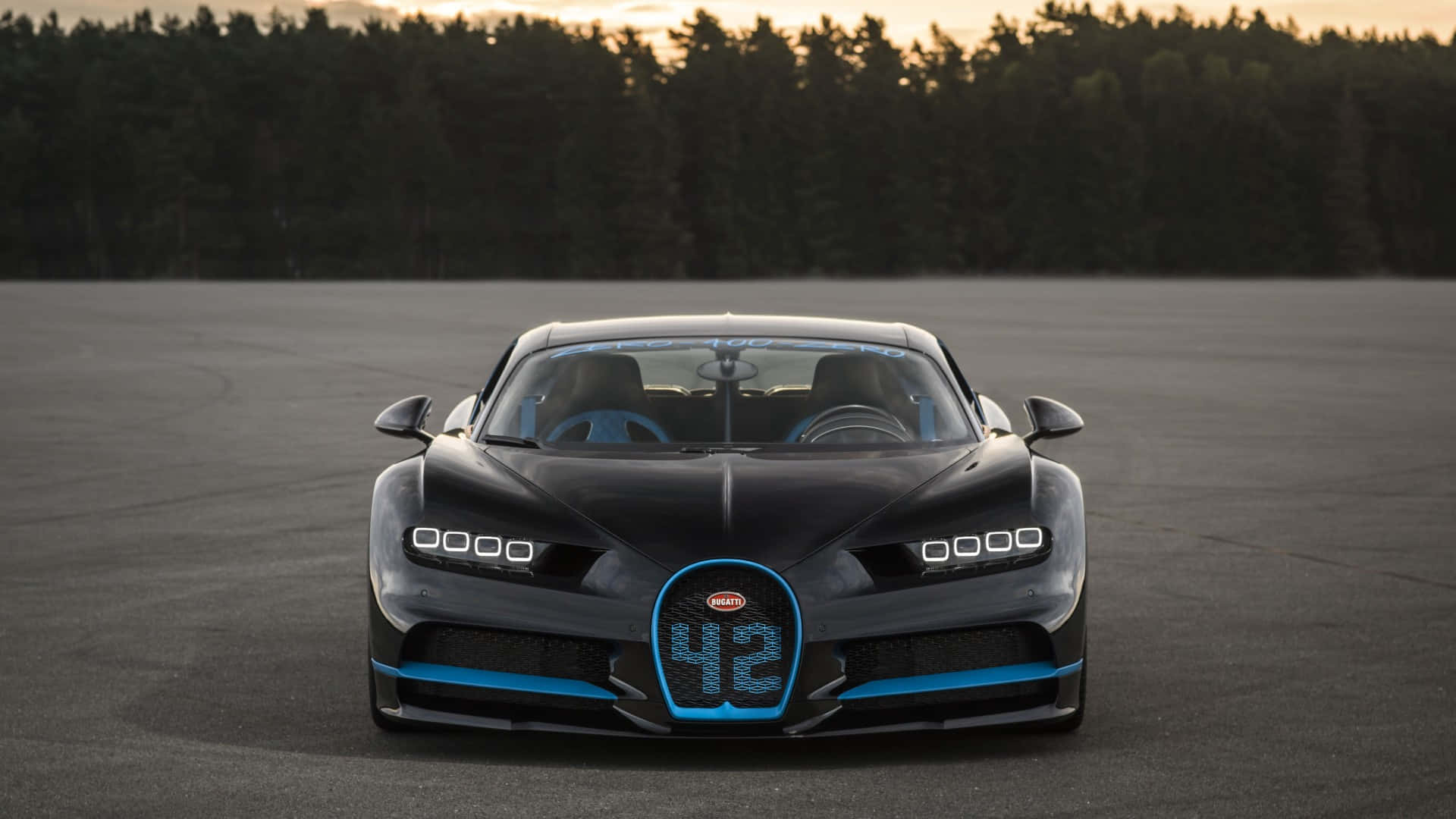 Bugatti Chiron 桌布 壁紙 — 一輛時尚的黑色Bugatti Chiron，帶有發光的藍色飾邊和車頭格柵上的42號，在日落時分停在空蕩的瀝青賽道上。