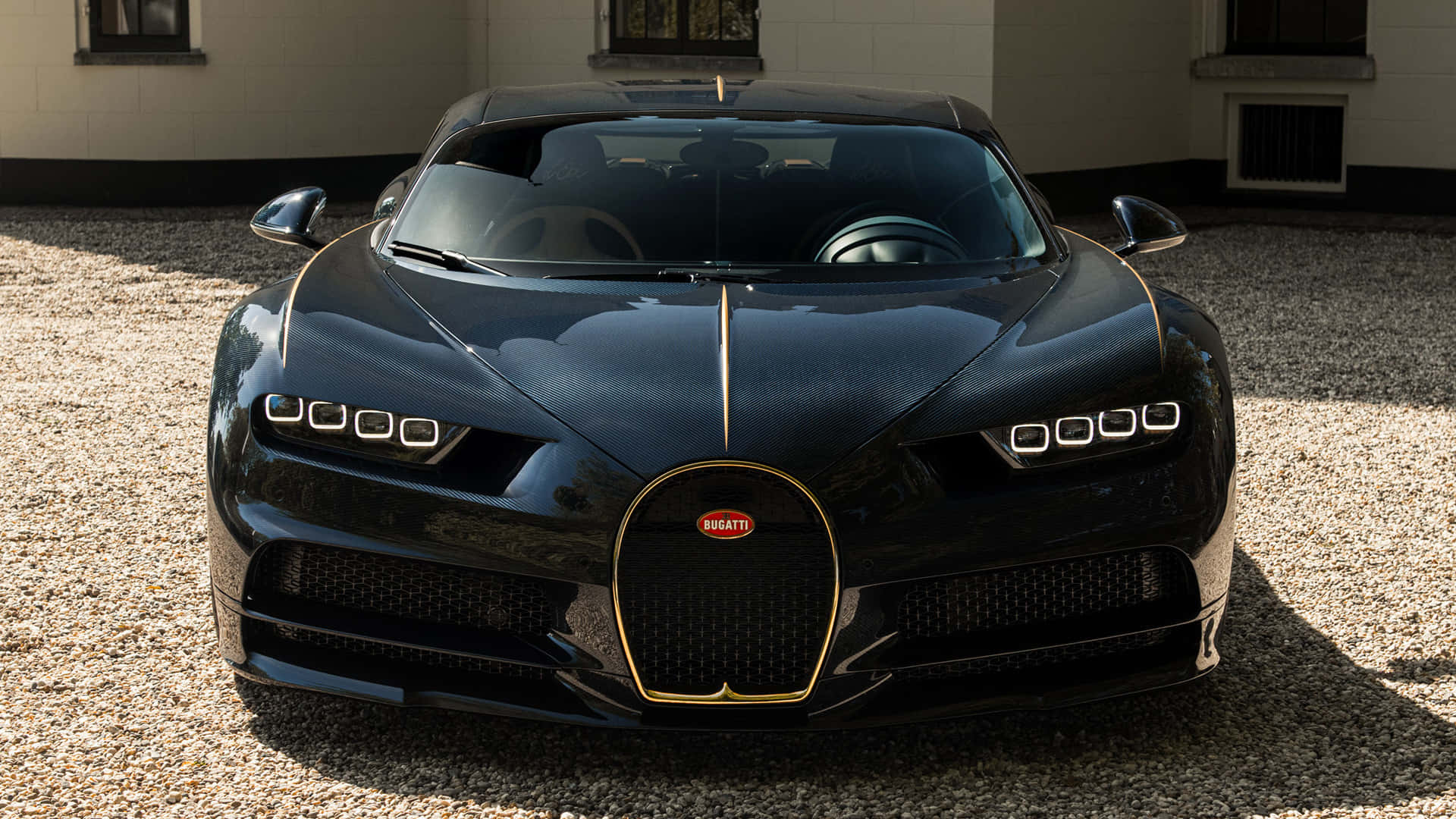 Bugatti Chiron 桌布 壁紙 — 一輛時尚的黑色Bugatti Chiron，帶有金色點綴，在陽光下於碎石車道上閃閃發光。