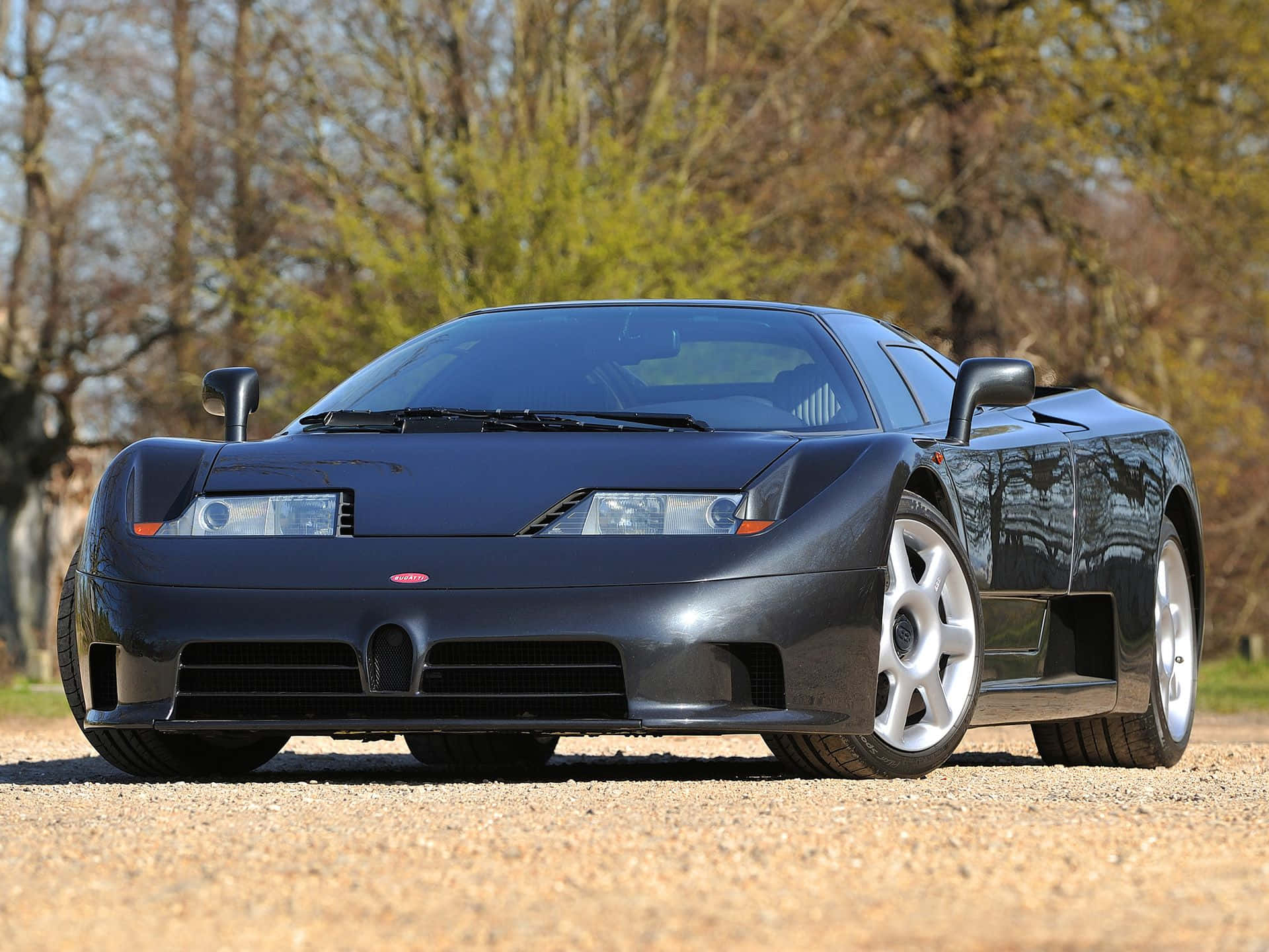 Bugatti Eb110 桌布 壁紙 — 一輛時尚的黑色布加迪 EB110 超級跑車在陽光明媚的碎石路上閃閃發光，周圍環繞著枝葉茂密的樹木。
