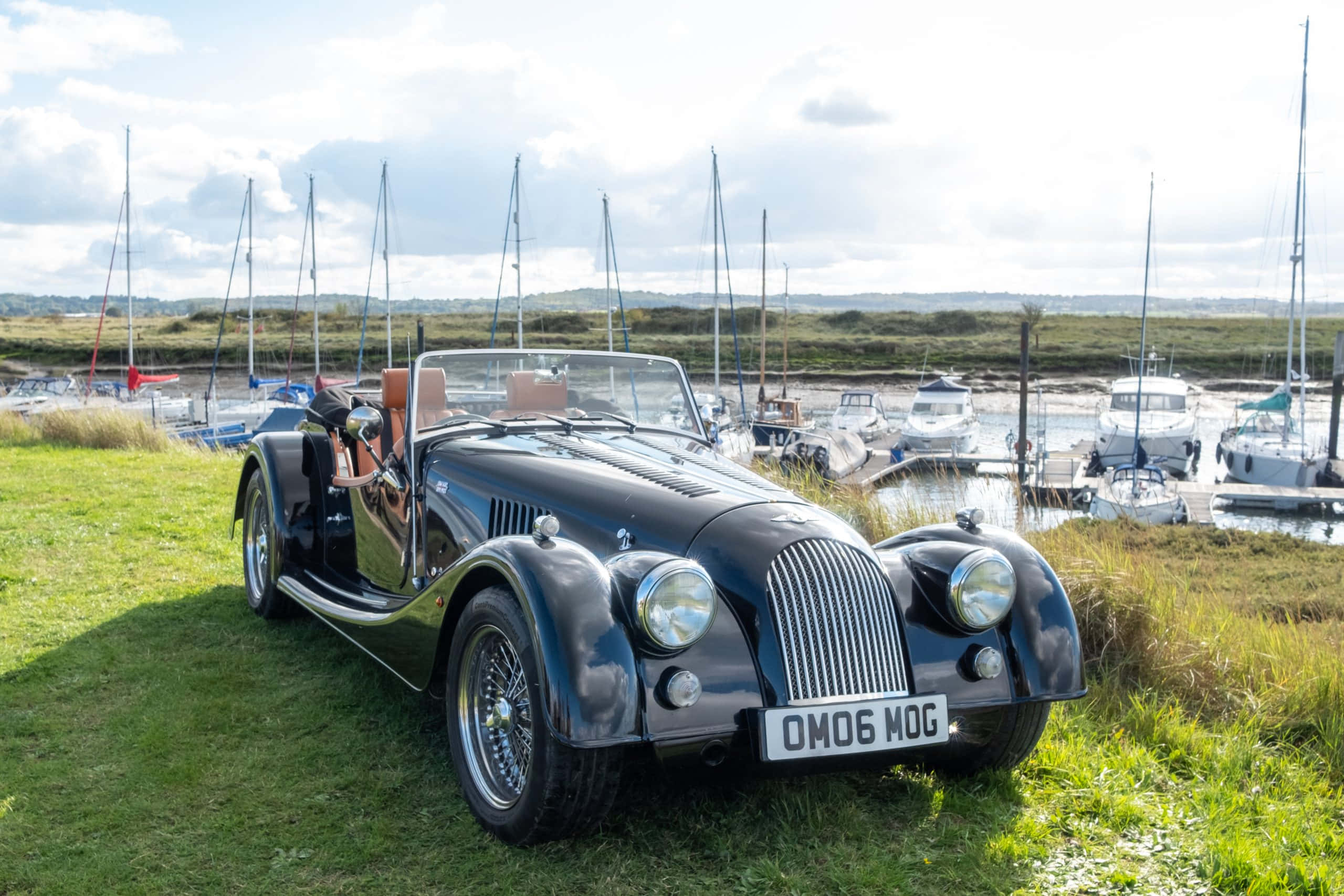 Morgan Roadster 桌布 壁紙 — 一架時尚的黑色經典敞篷跑車停泊在草地上，旁邊是停滿帆船的碼頭，天空局部多雲。