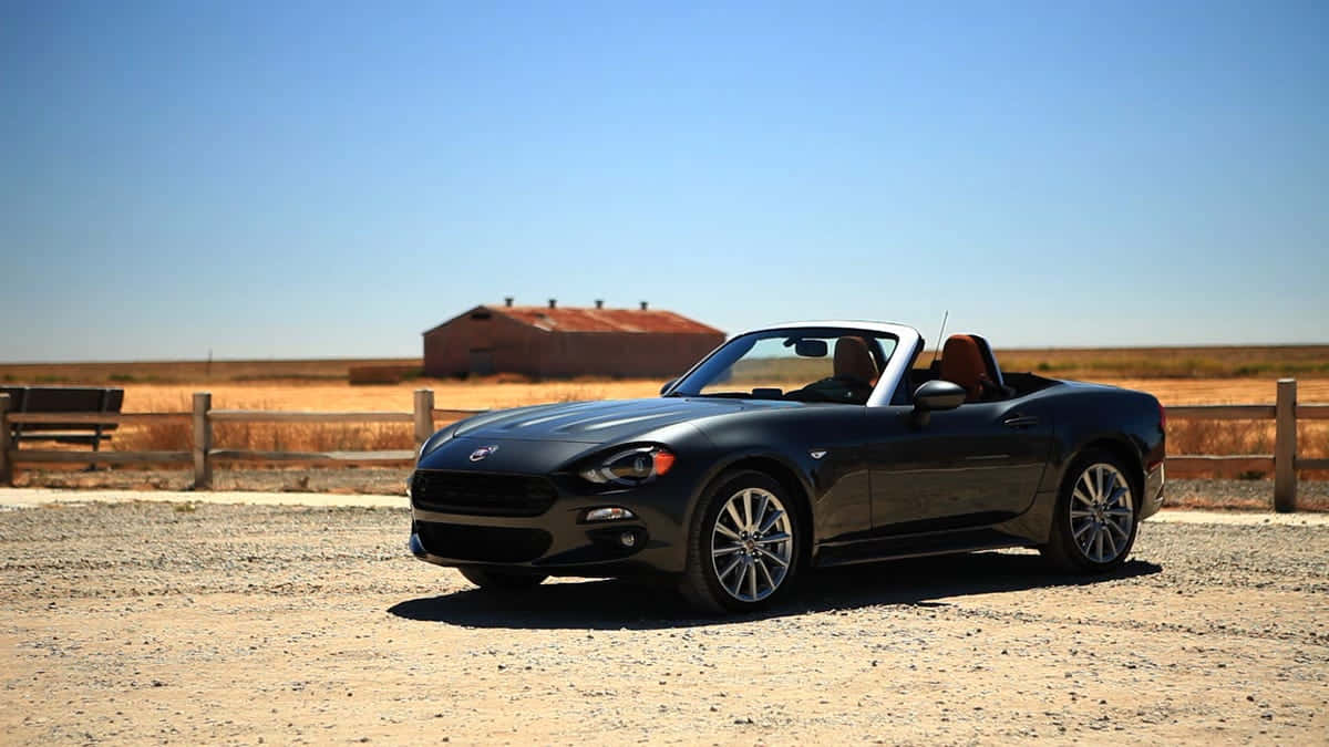 Fiat 124 Spider 桌布 壁紙 — 一架時尚黑色敞篷跑車停放在碎石地上，背景是晴朗藍天和一座質樸的紅色穀倉。