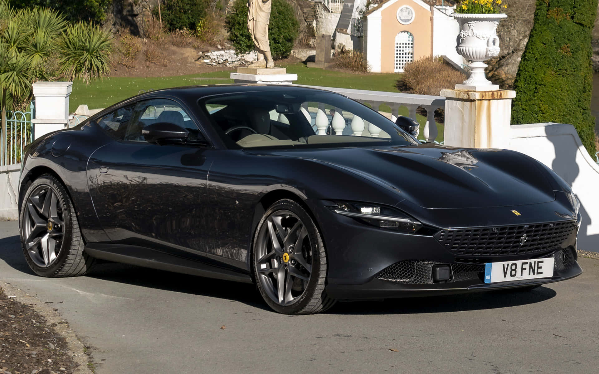 Ferrari Roma 桌布 壁紙 — 一輛時尚的黑色法拉利停泊在陽光普照的車道上，旁邊有白色圍欄和華麗的花園雕像。