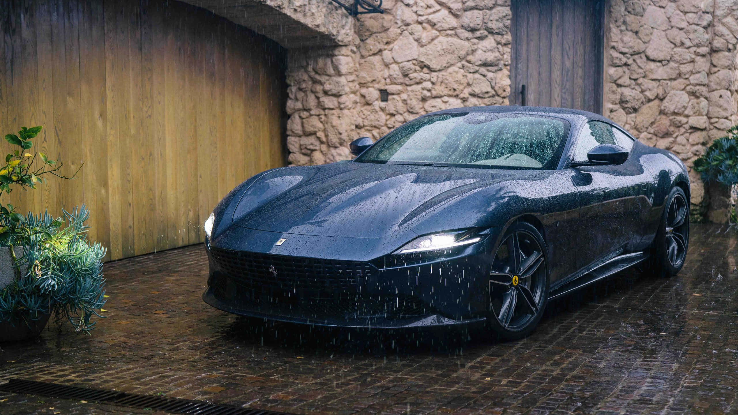 Ferrari Roma 桌布 壁紙 — 一架時尚的黑色法拉利跑車在鵝卵石庭院中，旁邊是一座質樸的石建築，在雨滴下閃閃發光。