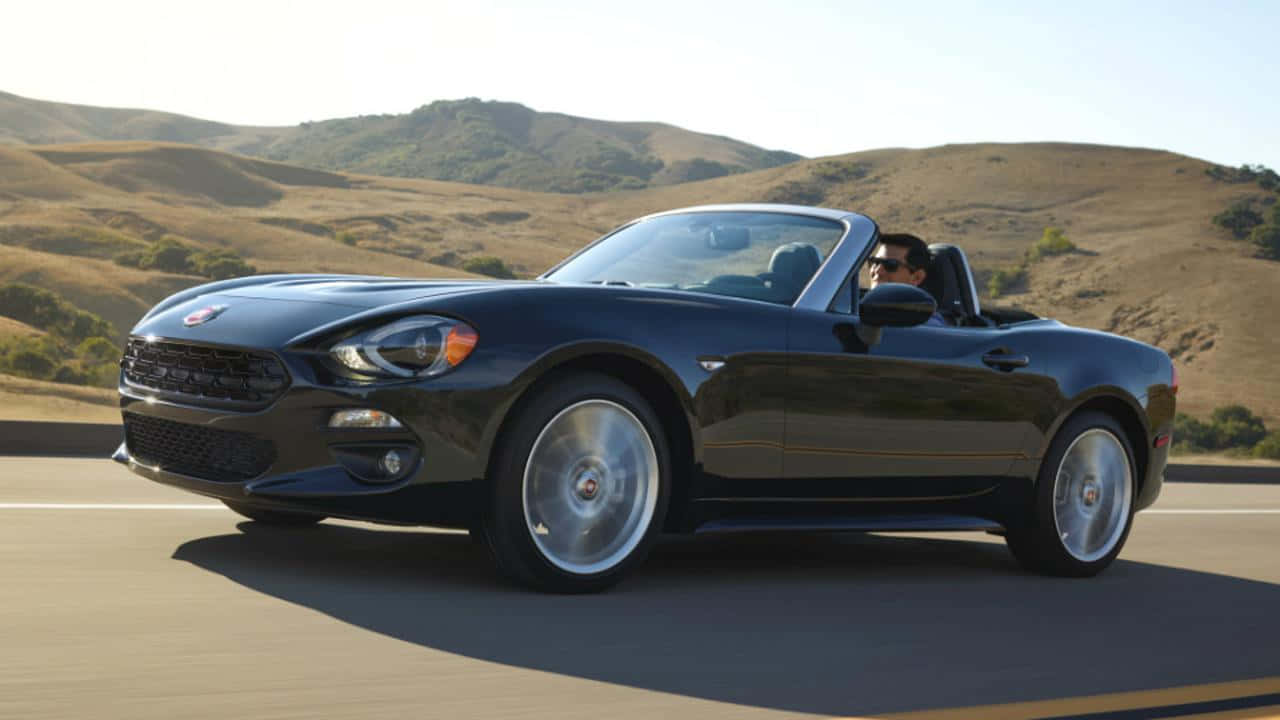 Fiat 124 Spider 桌布 壁紙 — 一台時尚的黑色Fiat 124 Spider敞篷車在陽光明媚的高速公路上巡航，背景是連綿的金色山丘。