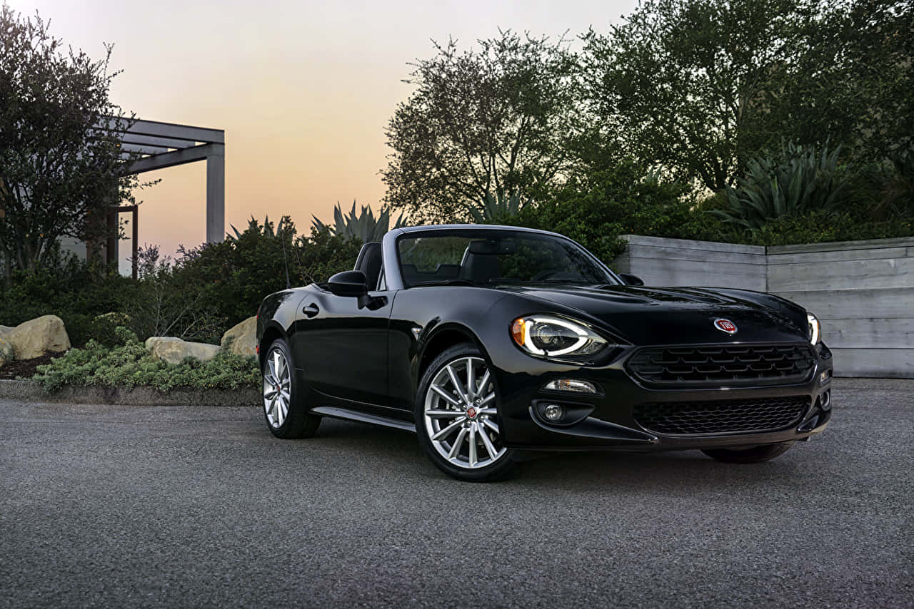 Fiat 124 Spider 桌布 壁紙 — 一架時尚的黑色Fiat 124 Spider敞篷車停放在鋪砌的車道上，背景是日落、樹木和一個現代化的涼棚。