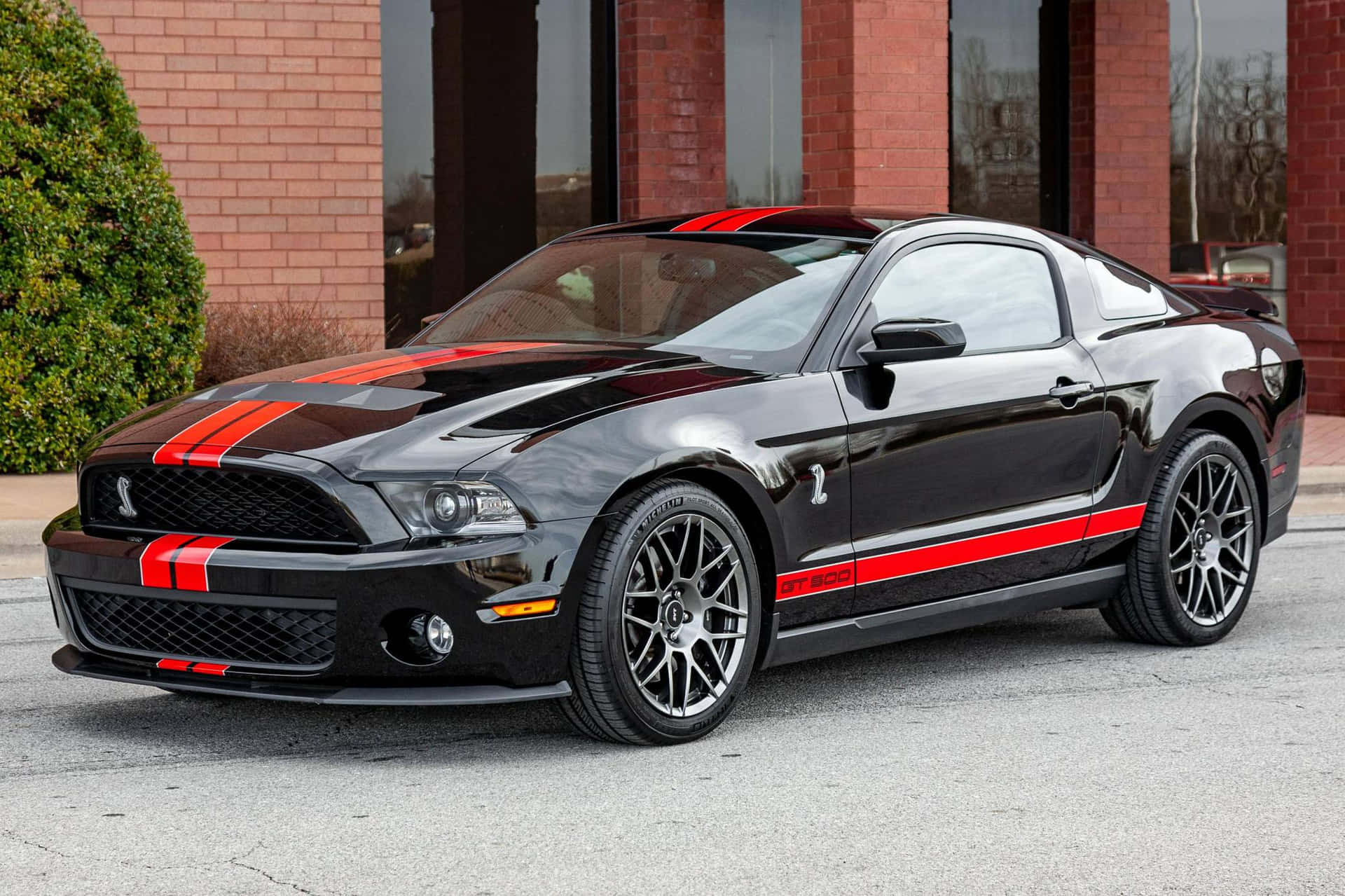  壁紙 — 一輛時尚的黑色福特野馬Shelby GT500，帶有醒目的紅色賽車條紋，停在磚砌建築前的瀝青路面上。
