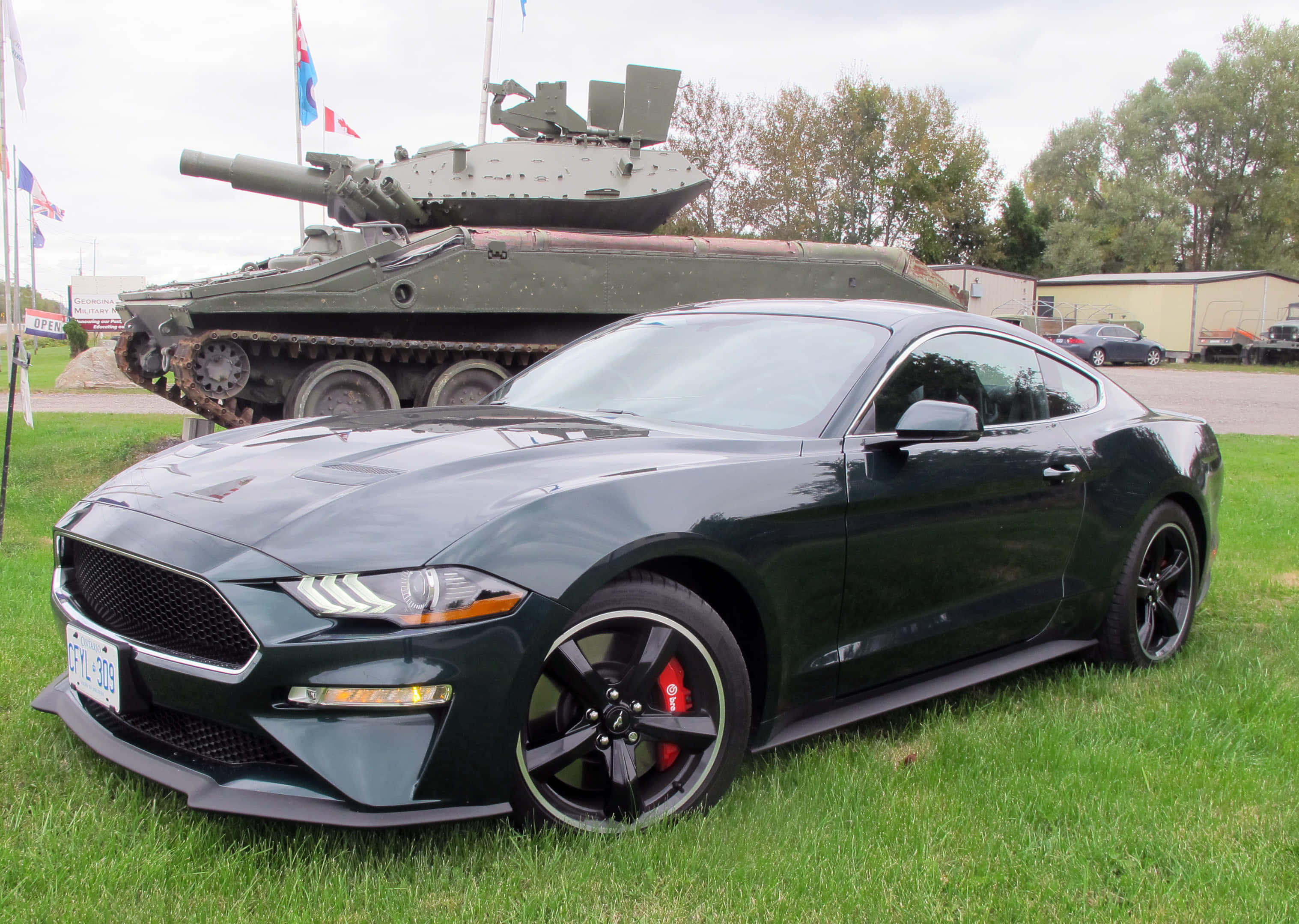 Ford Mustang Bullitt 桌布 壁紙 — 一架時尚的黑色福特野馬跑車停在草地上，背景有一輛軍用坦克，天空多雲。