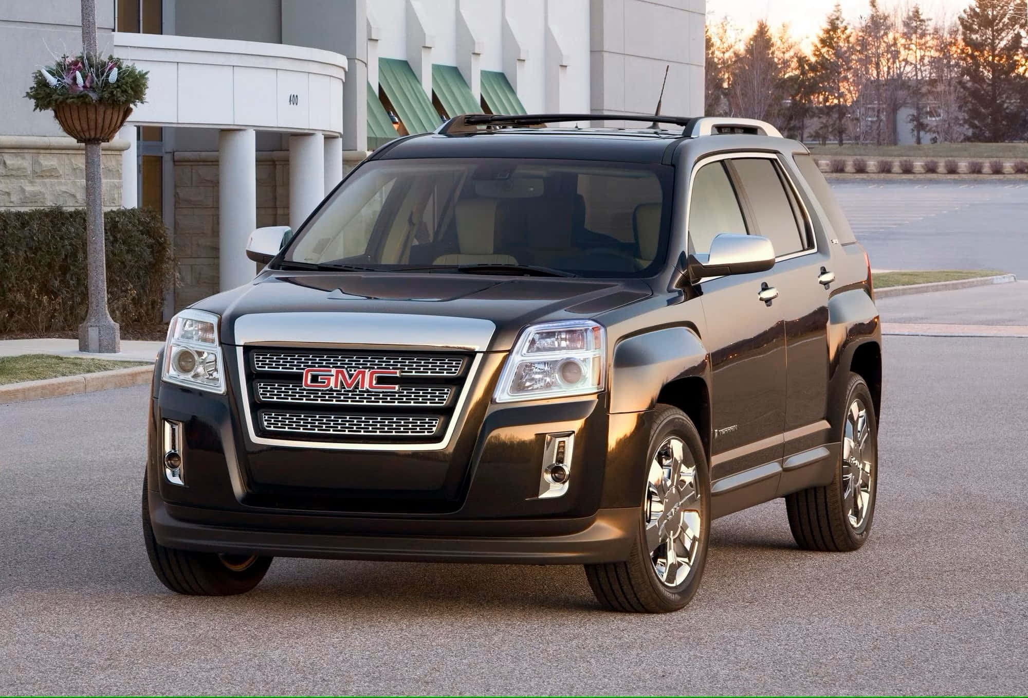 Gmc Terrain 桌布 壁紙 — 一輛時尚的黑色GMC Terrain SUV停放在鋪砌的停車場上，背景是現代建築和樹木，正值黃金時刻。
