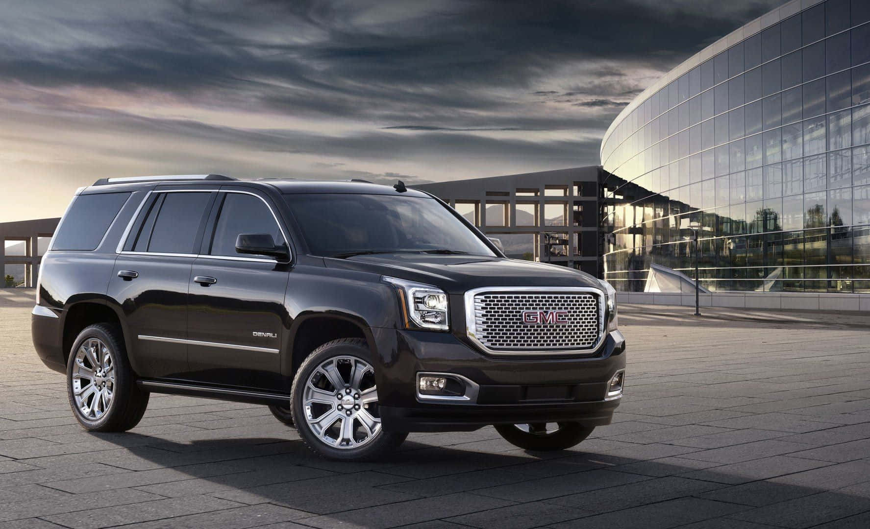 Gmc Yukon 桌布 壁紙 — 一輛時尚的黑色GMC Yukon SUV停放在現代玻璃建築前的瓷磚廣場上，天空佈滿戲劇性的雲層。