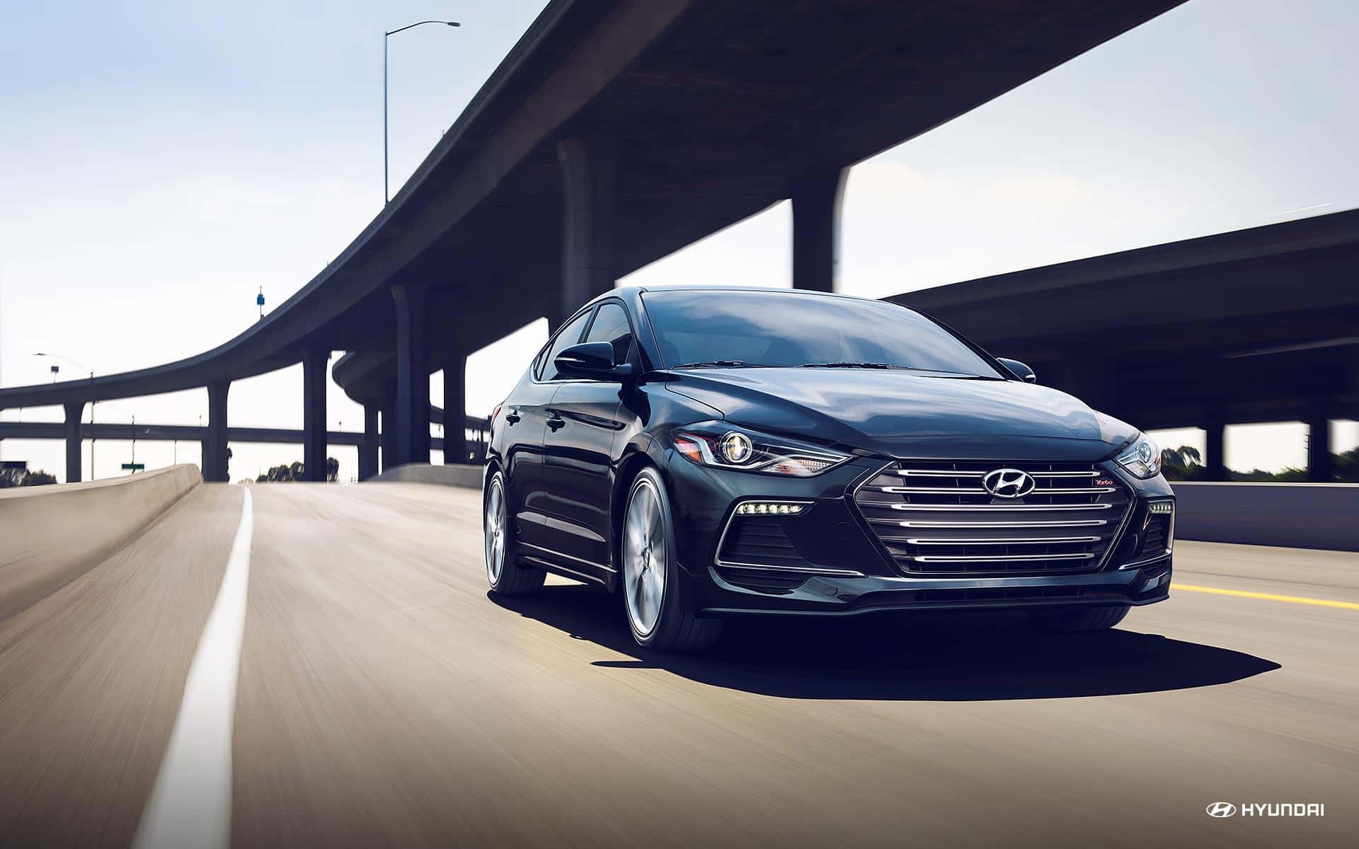 Hyundai Elantra 桌布 壁紙 — 一輛時尚的黑色現代轎車在陽光明媚的高速公路上疾馳，下方是雄偉的混凝土高架橋。