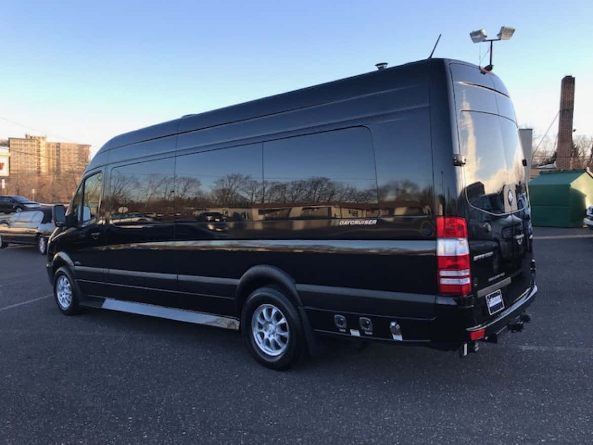 Mercedes Benz Sprinter 桌布 壁紙 — 一輛時尚的黑色豪華廂型車停在瀝青停車場上，天空清澈湛藍，光亮的車窗倒映著光禿的樹木。