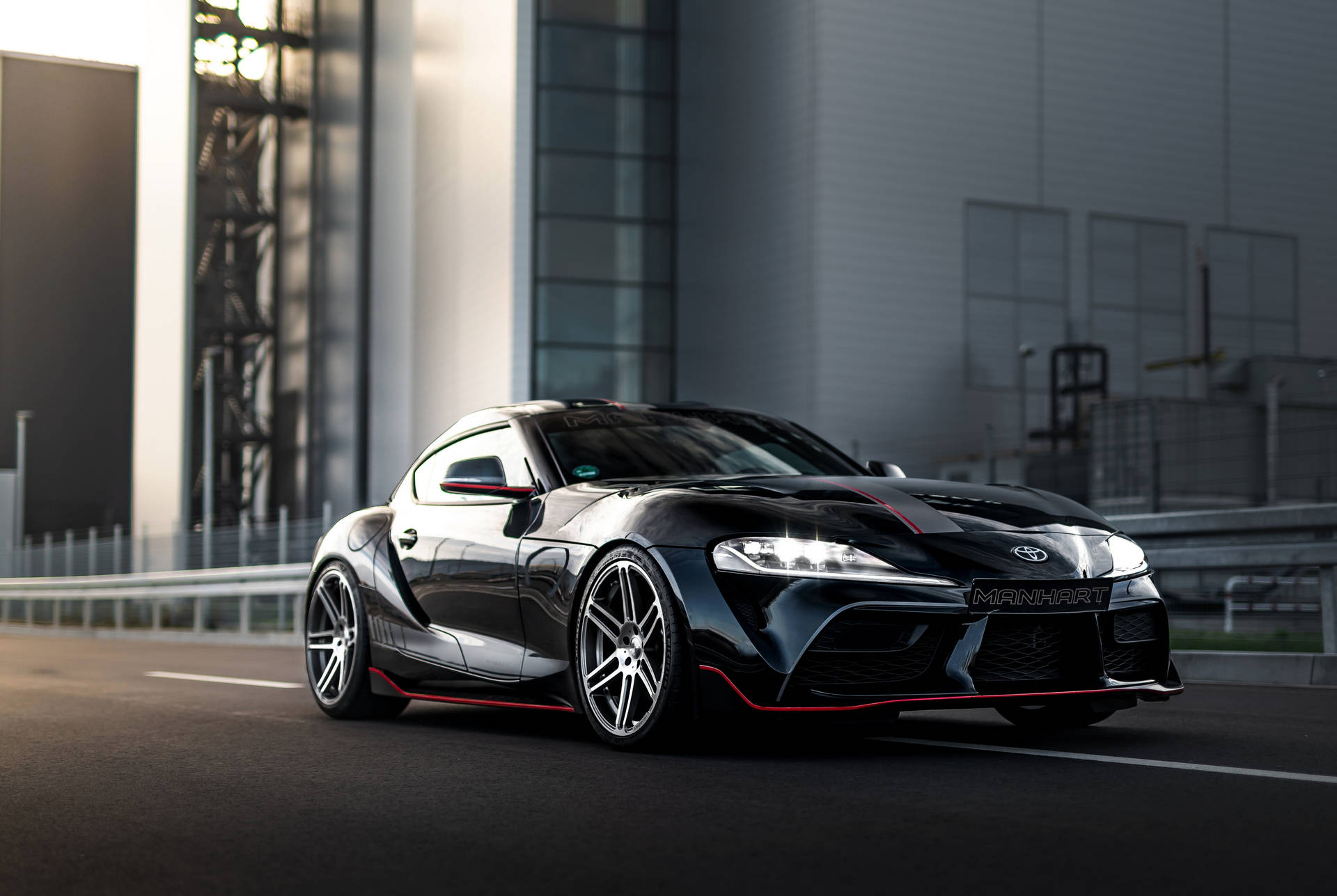 黑色Manhart豐田Supra，帶紅色飾邊，在工業建築附近的賽道上。 — Toyota Supra 壁紙