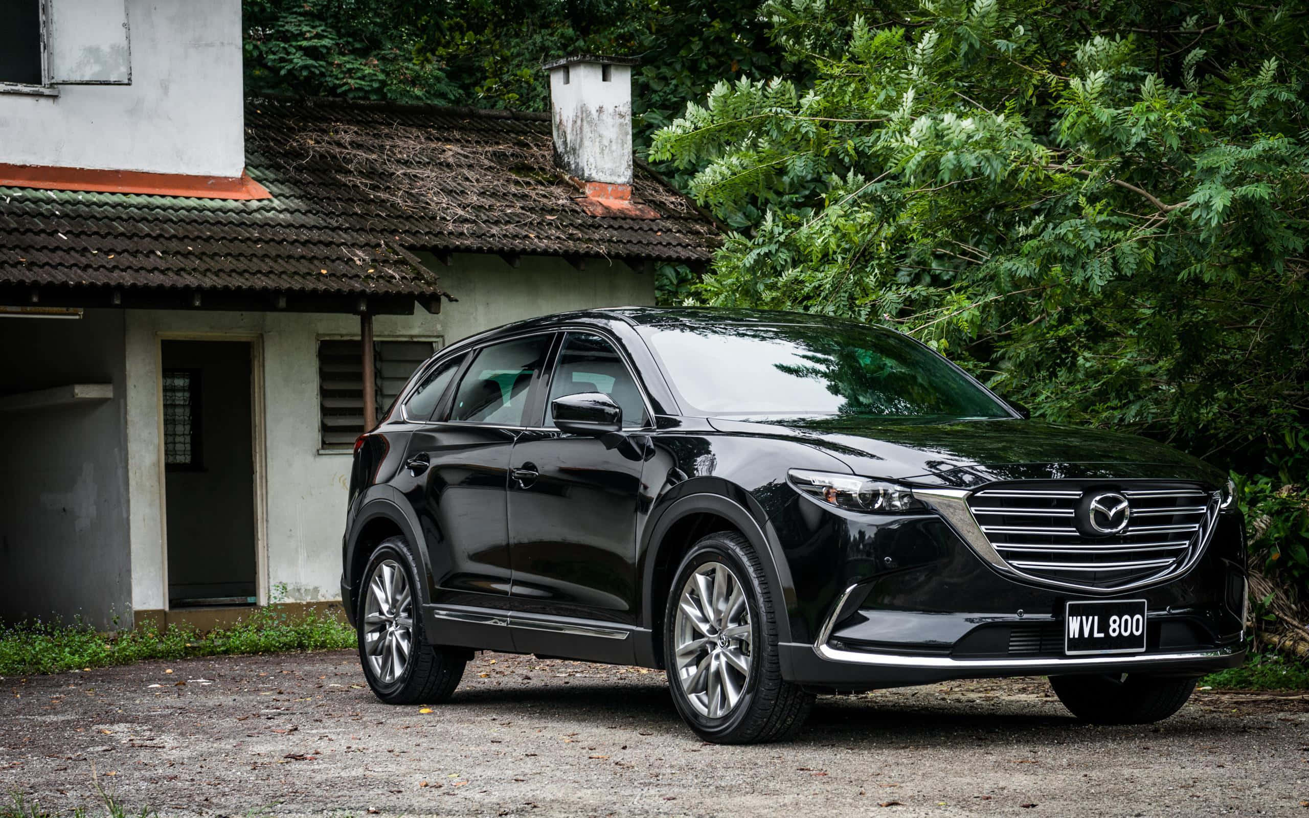 Mazda Cx-30 桌布 壁紙 — 一輛時尚的黑色Mazda CX-9 SUV停泊在碎石路上，前面是一座被茂密綠葉環繞的質樸房屋。