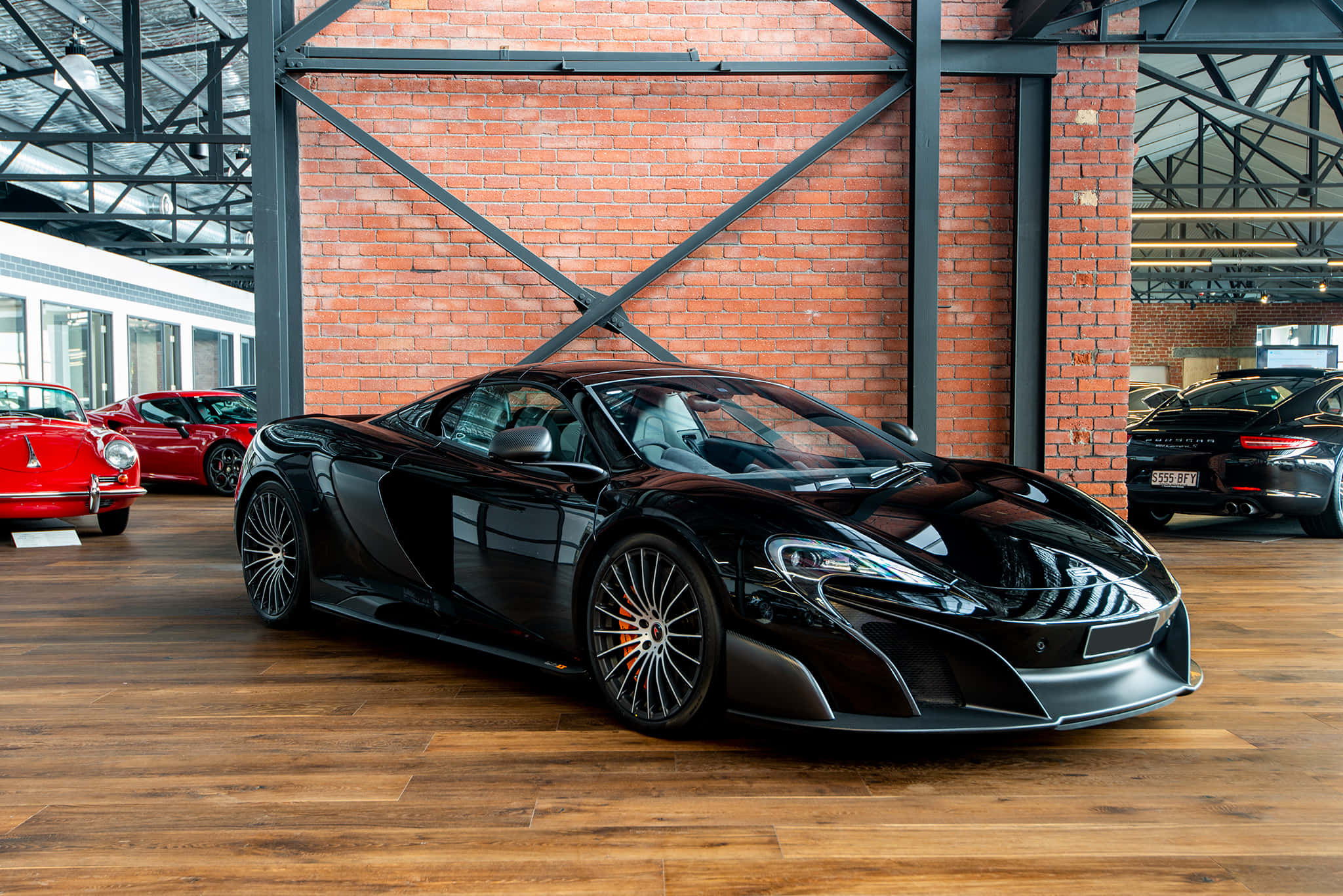 Mclaren 675lt Spider 桌布 壁紙 — 一輛時尚的黑色McLaren跑車在明亮的燈光下，在寬敞的磚牆和木地板的車庫中閃閃發光，旁邊還有其他豪華汽車。