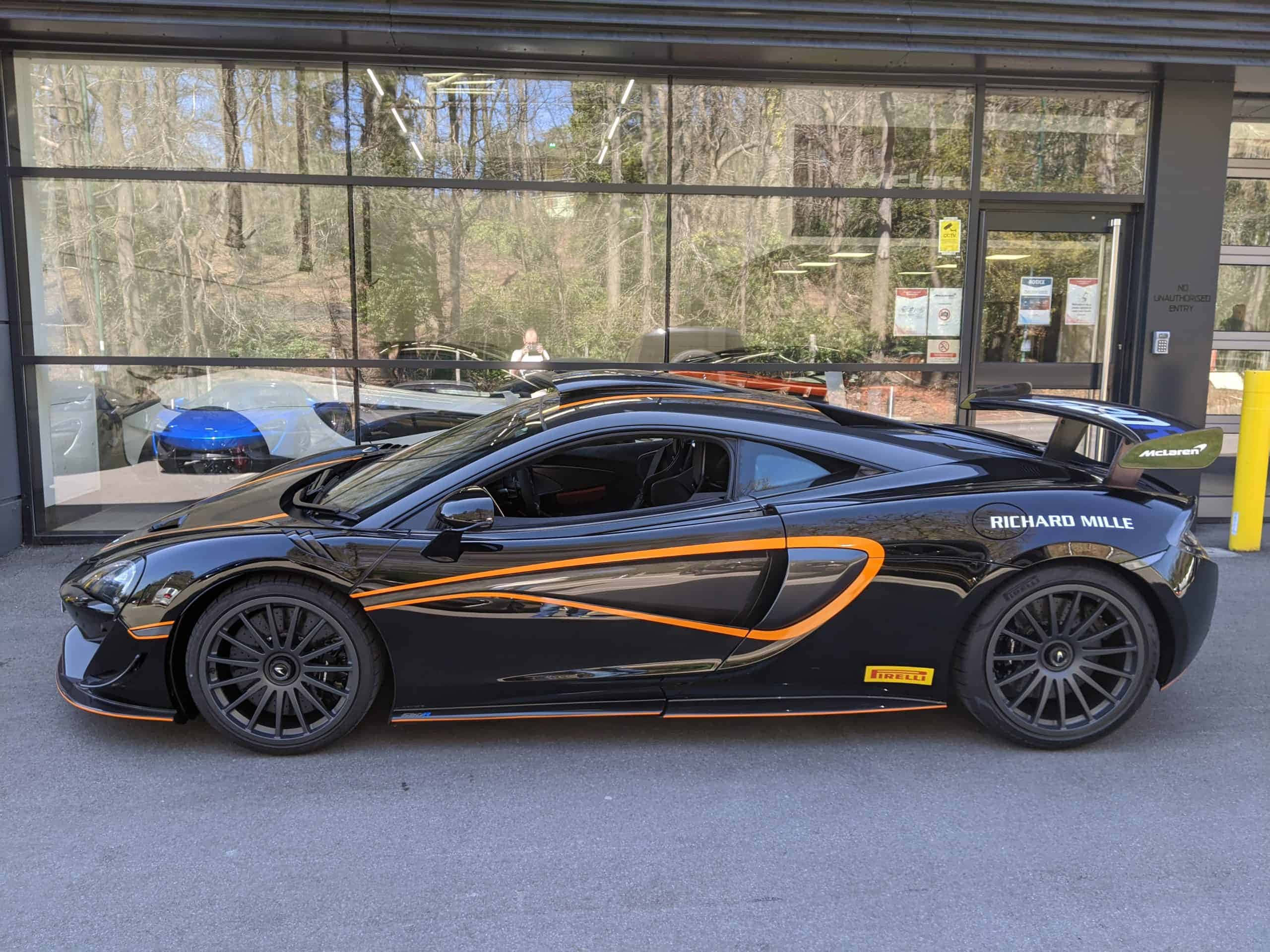Mclaren 620r 桌布 壁紙 — 一輛時尚的黑色McLaren跑車帶有橙色點綴，停在玻璃門面的經銷商外。