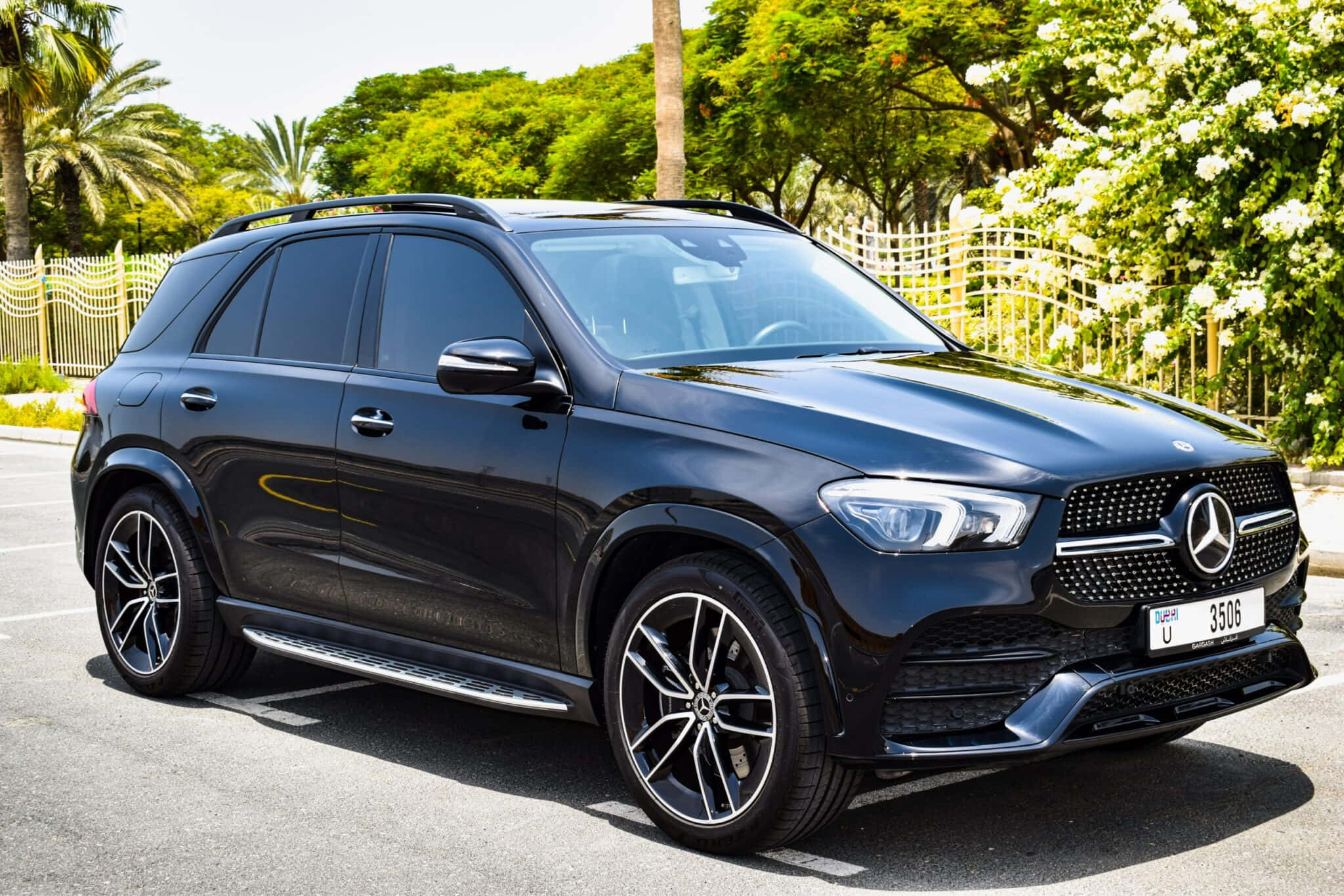 Mercedes Benz Gle-class 桌布 壁紙 — 一輛時尚的黑色賓士 GLE SUV 停放在陽光普照的停車場，周圍環繞著茂密的綠樹和白色開花灌木。