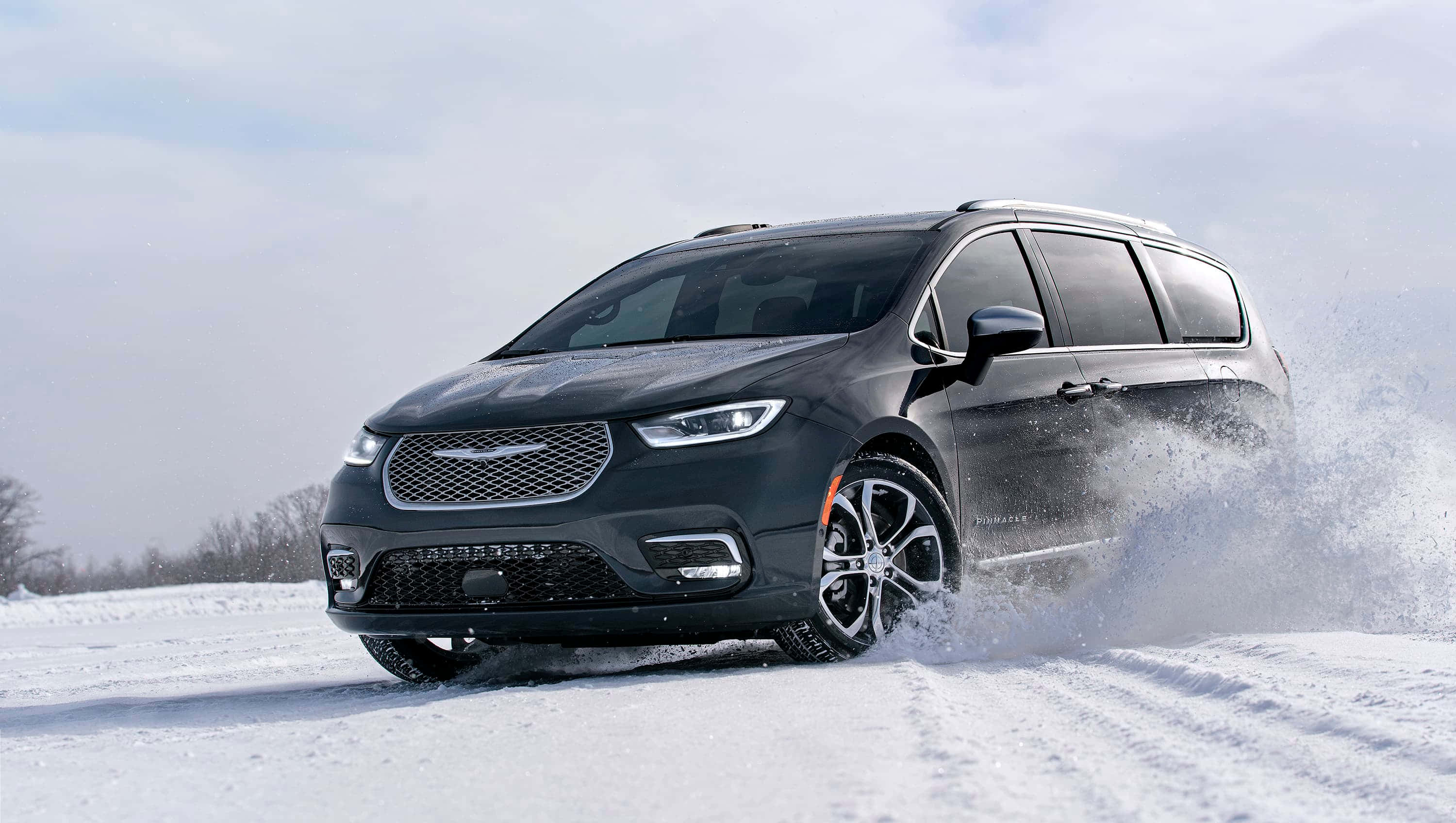 Chrysler Pacifica 桌布 壁紙 — 一輛時尚的黑色小型貨車在深雪中行駛，揚起白色雪花，映襯著蒼白的冬日天空。