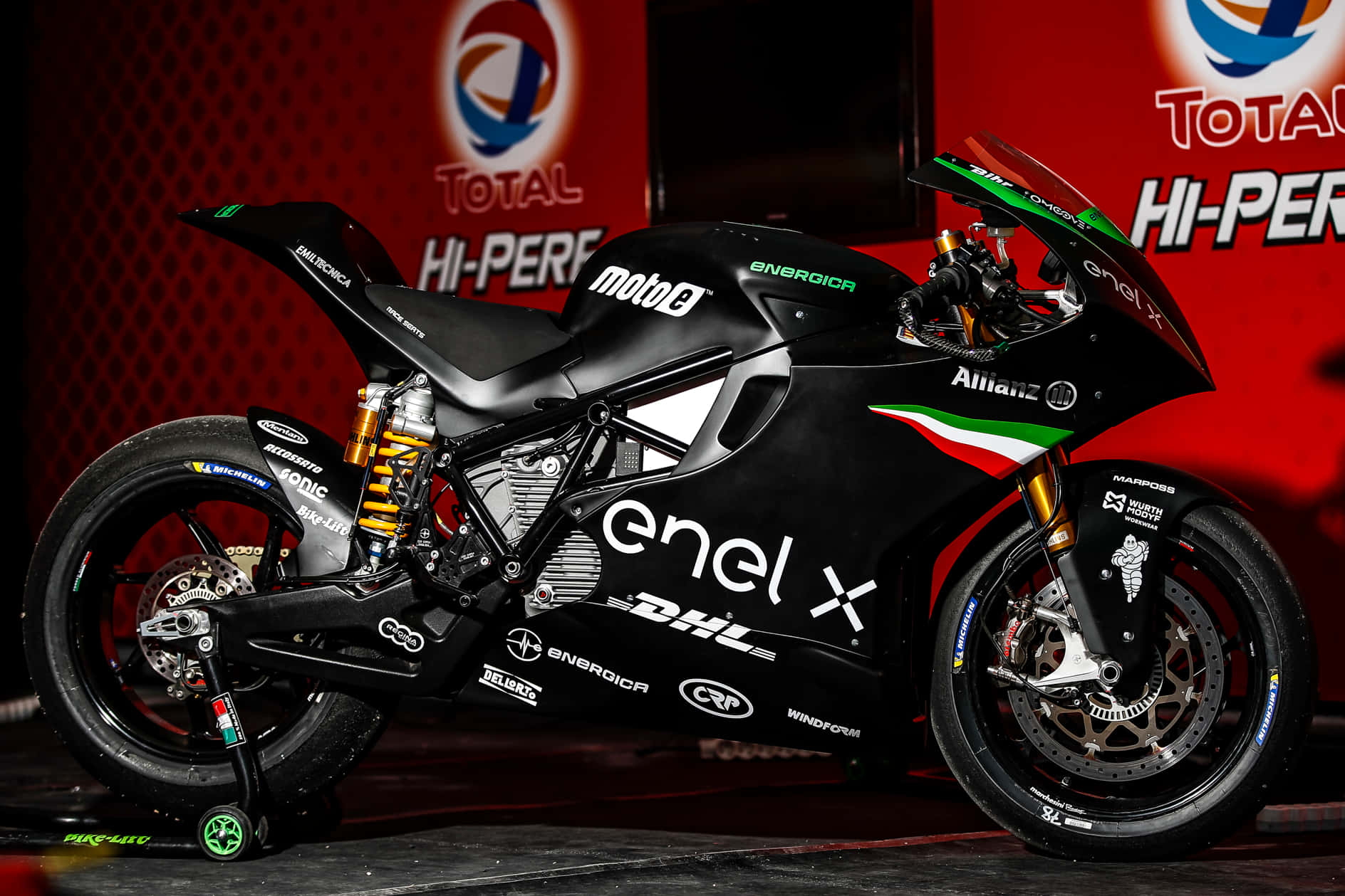  壁紙 — 一輛時尚的黑色Moto2賽車摩托車，帶有綠色點綴和贊助商標誌，在紅色品牌背景的舞台上展示。