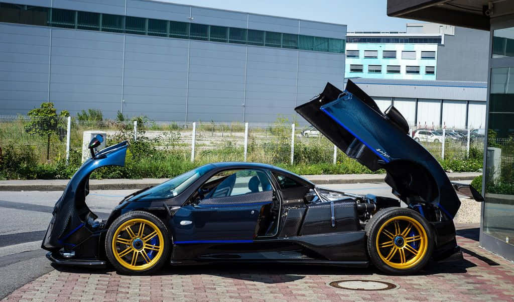 Pagani Zonda Tricolore 桌布 壁紙 — 一輛時尚的黑色帕加尼超級跑車，配備金色輪圈和打開的鷗翼門，停在磚砌車道上，在明亮的日光下。
