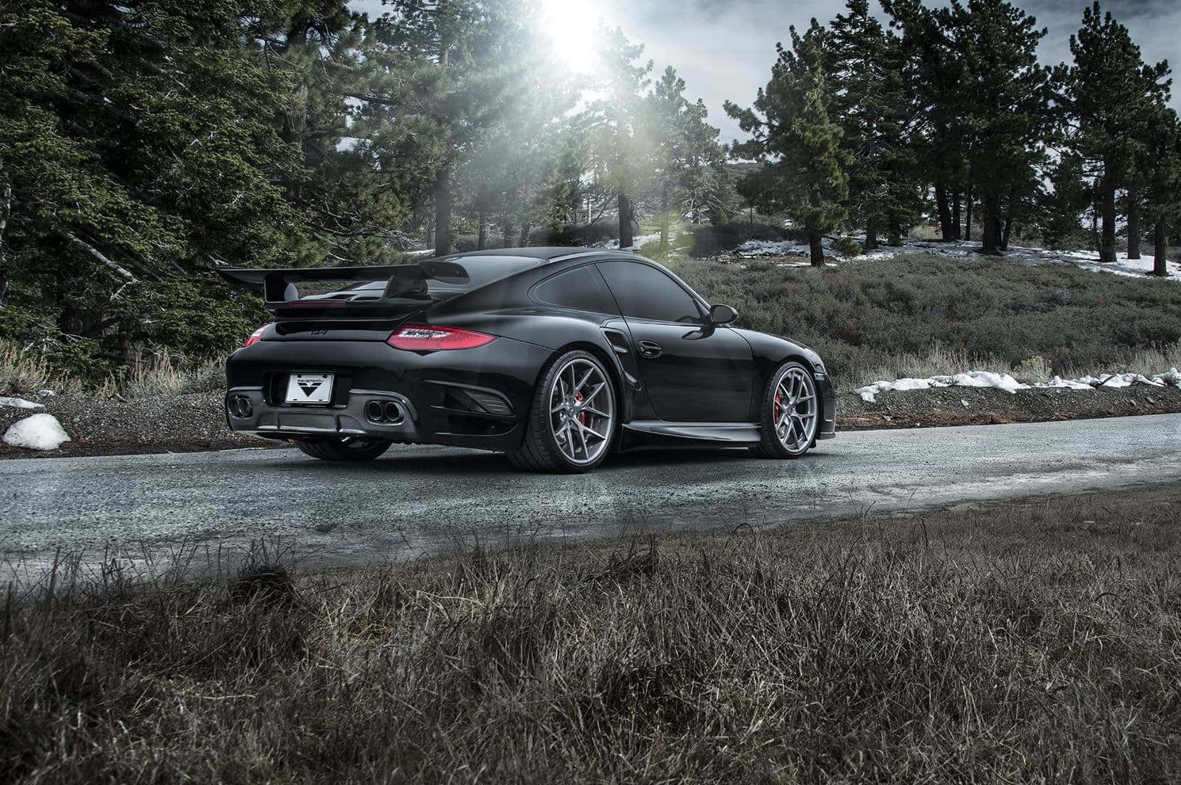 Porsche 997 桌布 壁紙 — 一輛時尚的黑色保時捷跑車，帶有大型後擾流板，停放在森林道路上，陽光穿過松樹照射下來。