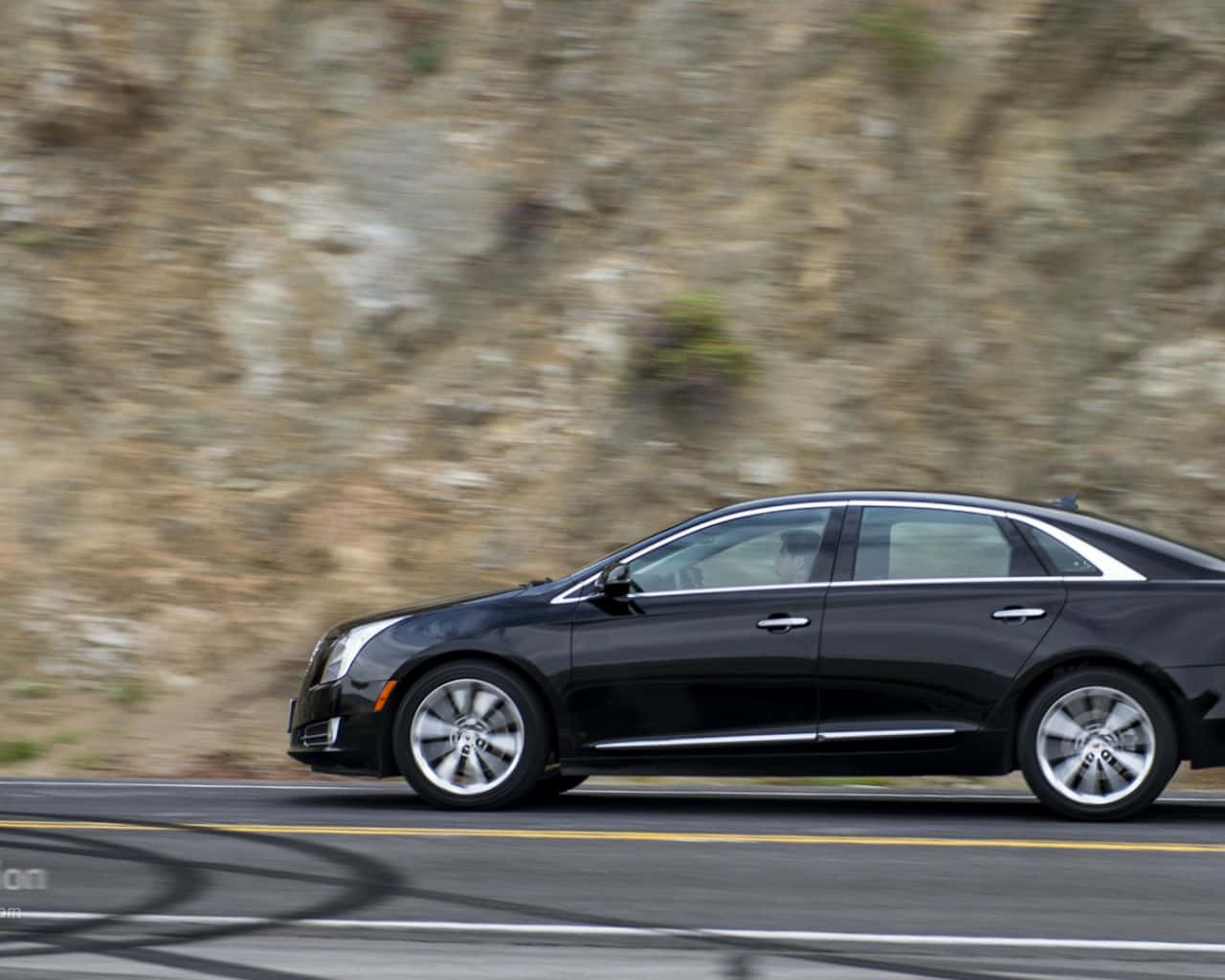 Cadillac Xts 桌布 壁紙 — 一輛時尚黑色轎車沿著蜿蜒的道路疾馳，背景是模糊的岩石懸崖。