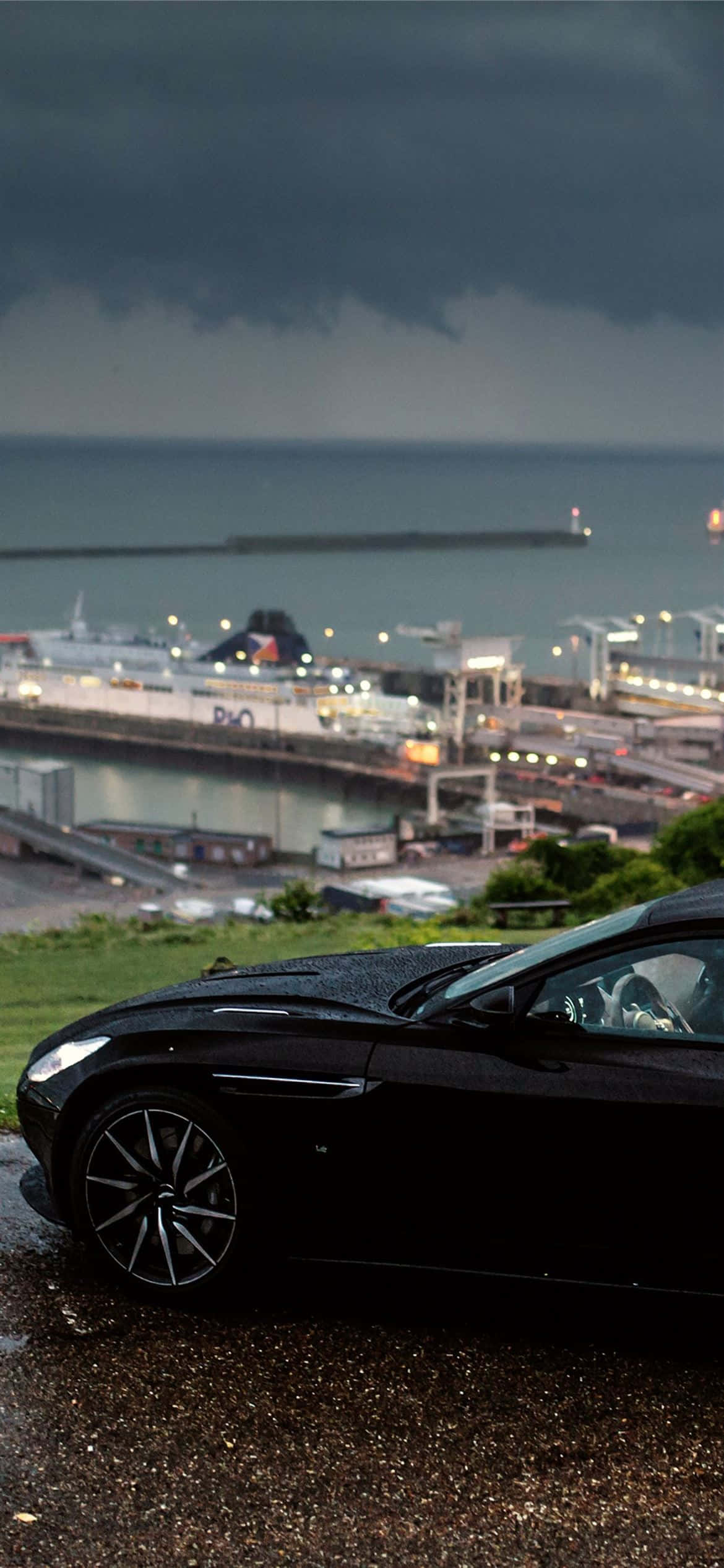 Aston Martin Db11 桌布 壁紙 — 一輛時尚的黑色跑車停在濕潤的山丘上，俯瞰著風暴中的港口，港口燈光閃爍。