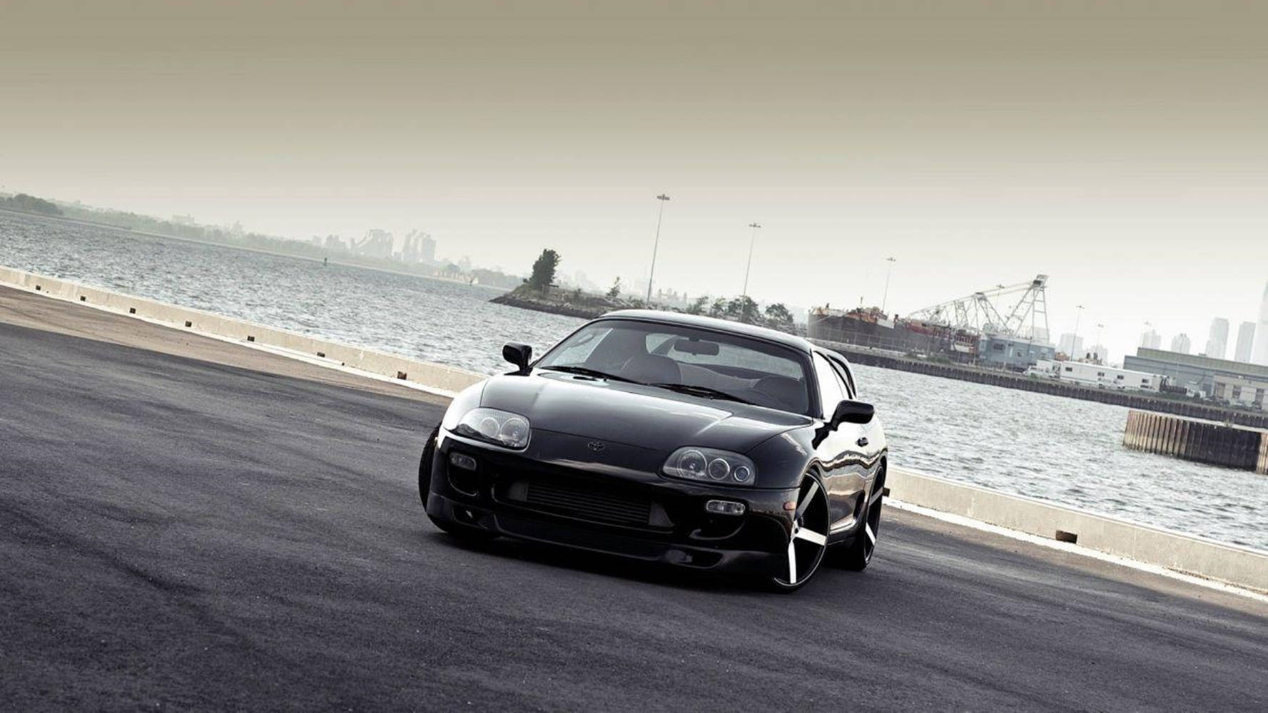 黑色跑車在工業港口附近的海濱道路上行駛。 — Toyota Supra 壁紙