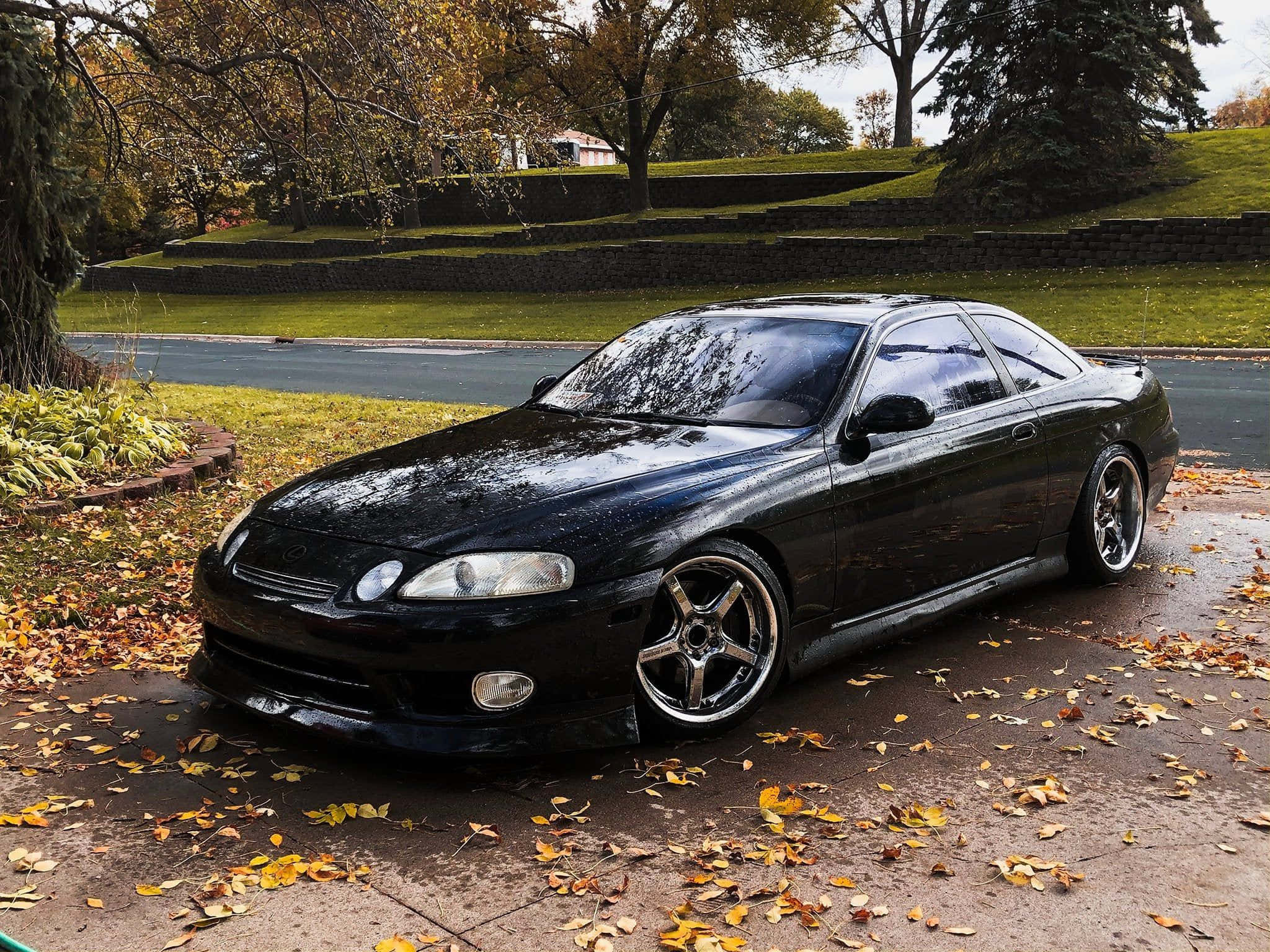 Lexus Sc 300 桌布 壁紙 — 一輛時尚黑色雙門跑車停泊在落葉滿佈的車道上，背景是秋天的樹木和長滿草的小山丘。