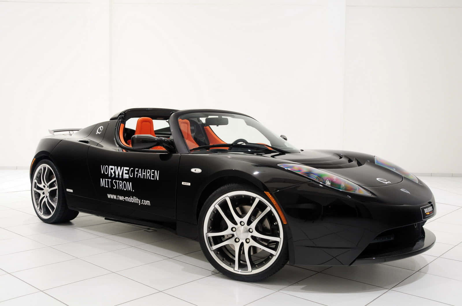 Tesla Roadster 桌布 壁紙 — 一架時尚的黑色Tesla Roadster敞篷車，配橙色座椅和白色輪圈，停放在明亮的白色瓷磚工作室中。