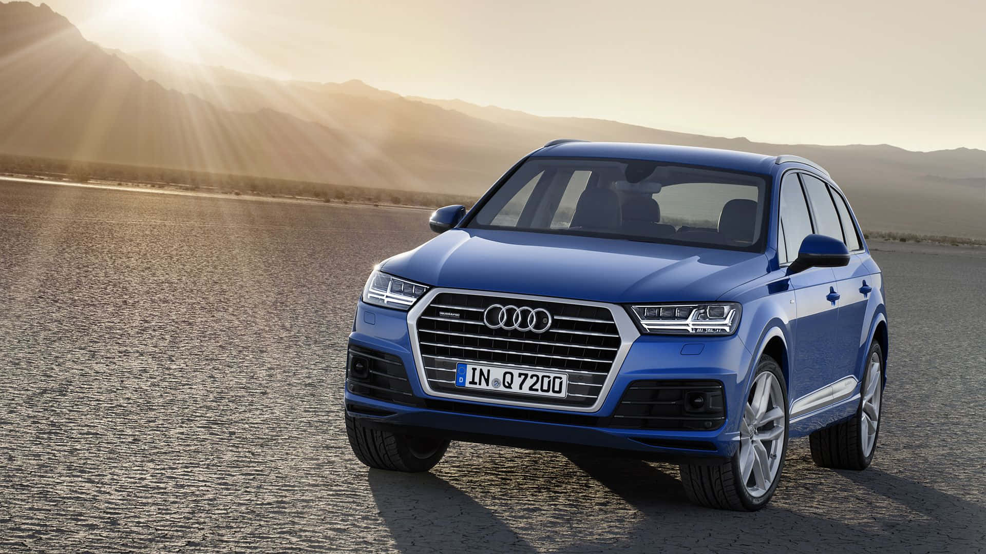 Audi Q7 桌布 壁紙 — 一輛時尚的藍色奧迪Q7 SUV停泊在陽光明媚的沙漠公路上，背景有山脈和金色光芒。
