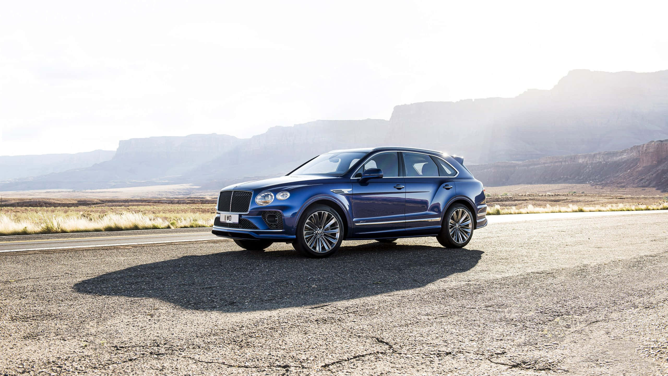 Bentley Bentayga 桌布 壁紙 — 一輛時尚的藍色Bentley Bentayga SUV停放在沙漠公路上，背景是崎嶇的山脈，在明亮的日光下。