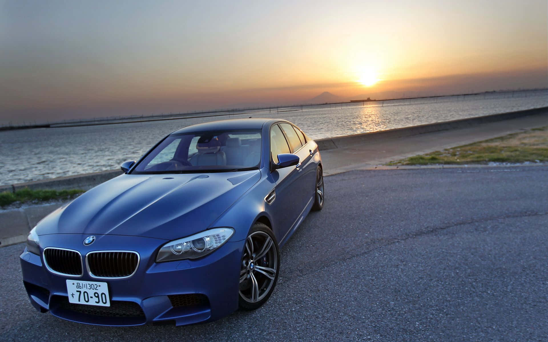 Bmw M5 桌布 壁紙 — 一輛時尚的藍色BMW M5停在海濱道路上，夕陽在水面上投下金色光芒。