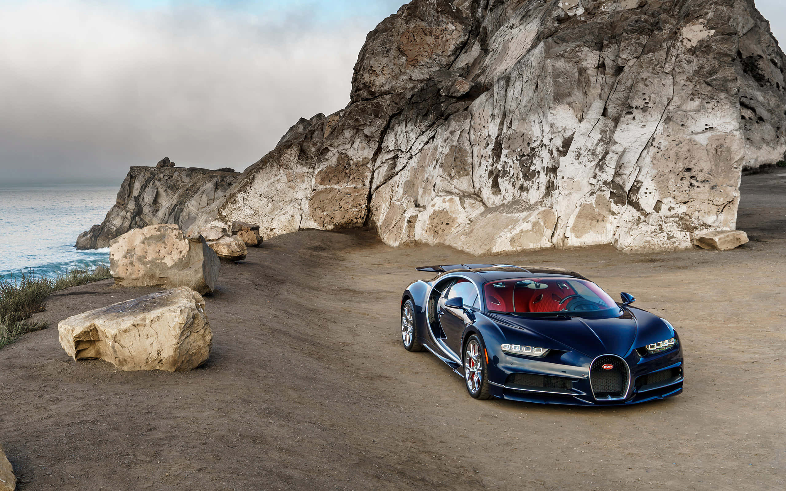 Bugatti Chiron 桌布 壁紙 — 一架時尚的藍色Bugatti Chiron停泊在崎嶇的海岸懸崖上，伴隨著洶湧的海浪和戲劇性的岩石 formations，天空多雲。
