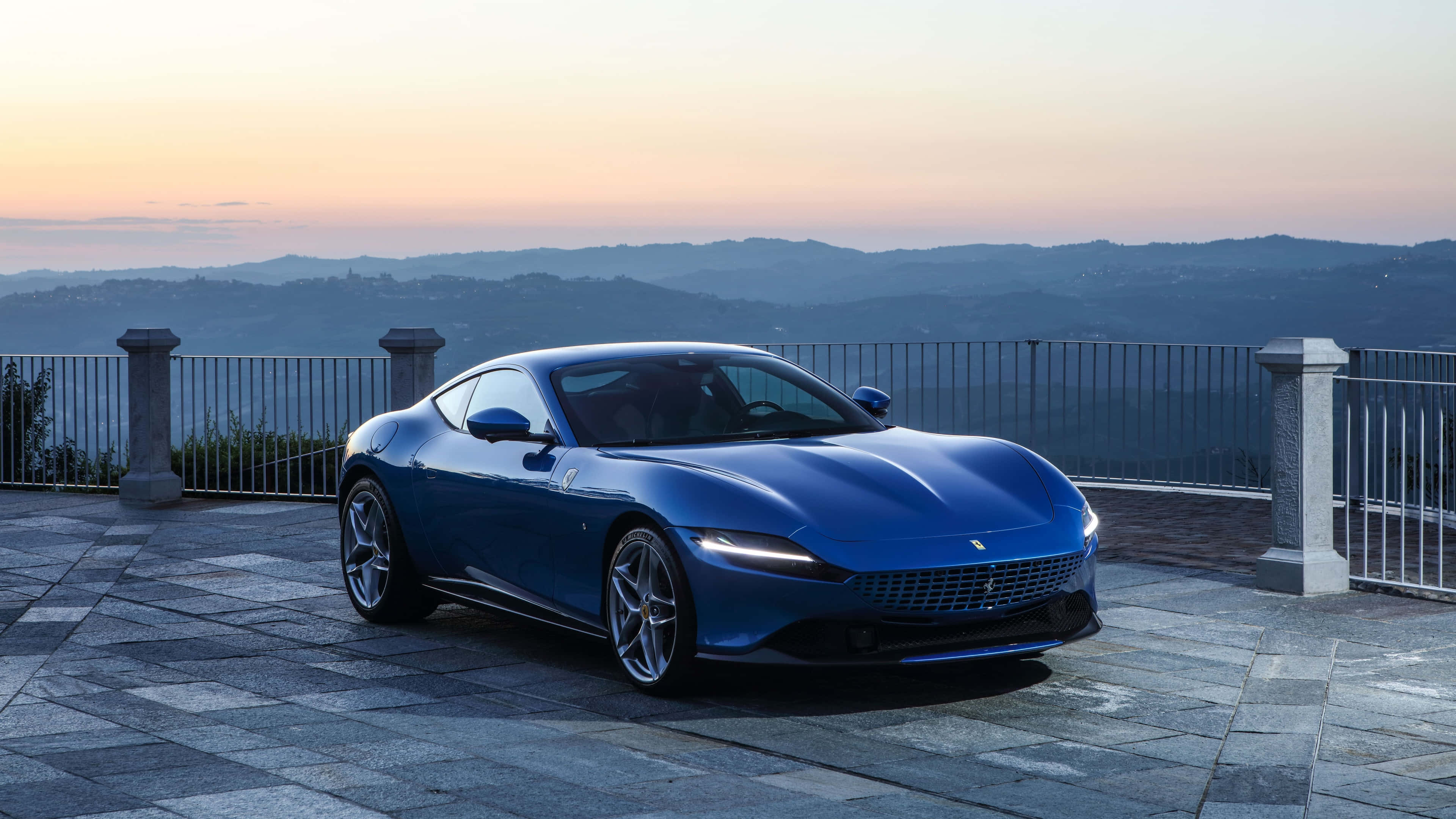 Ferrari Roma 桌布 壁紙 — 一架時尚的藍色法拉利跑車停泊在石頭露台上，俯瞰日落時分的連綿山丘。
