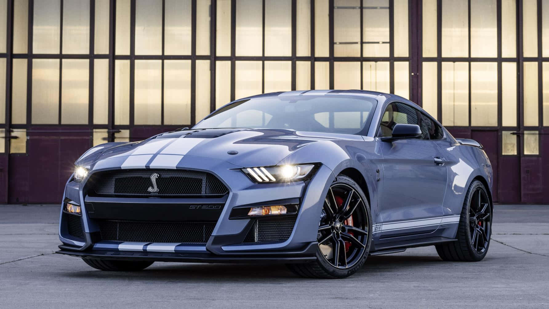  壁紙 — 一輛時尚的藍色福特野馬Shelby GT500，帶有白色賽車條紋和發光頭燈，停在一座現代玻璃和金屬建築前。