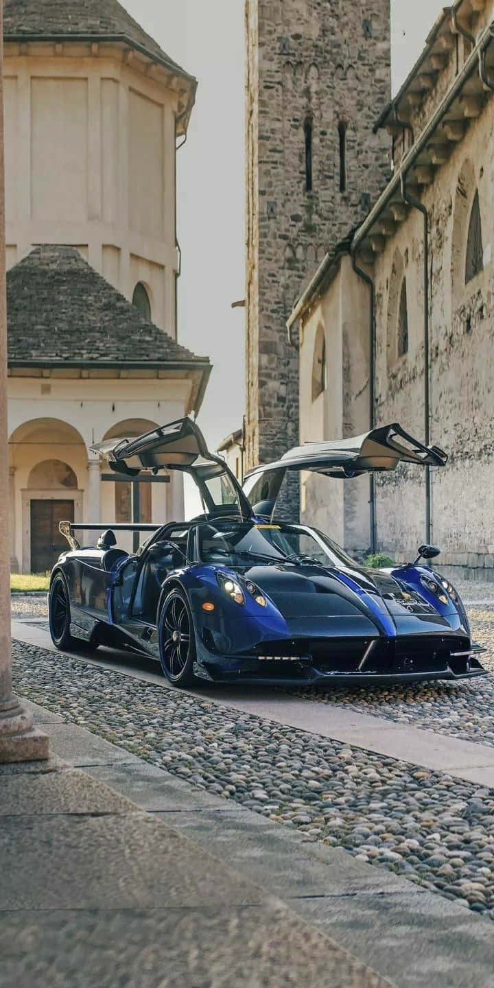 Pagani Zonda Tricolore 桌布 壁紙 — 一架時尚的藍色超跑，配備鷗翼車門，停在鵝卵石街道上，旁邊是歷史悠久的石建築。